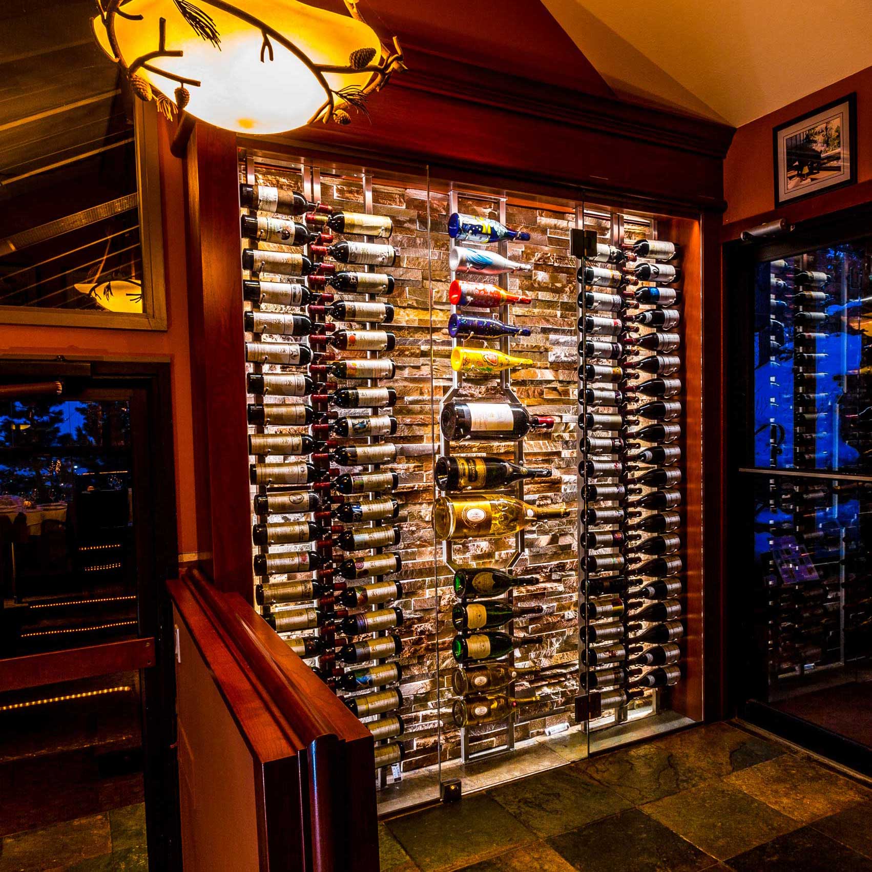 The image shows a well-lit wine rack display in a room with warm lighting. The rack is filled with numerous bottles of wine, organized horizontally on metal shelves against a brick wall. The setting appears to be elegant, possibly in a restaurant or a wine cellar. A decorative ceiling light fixture is visible above, and there is a framed picture on the wall to the right.