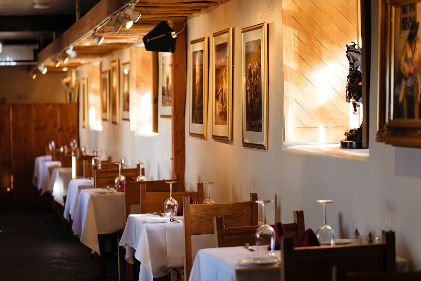 The image shows the interior of a cozy restaurant with wooden furniture. There are several tables lined up along the wall, each covered with white tablecloths and set with wine glasses and cutlery. The walls are adorned with framed artwork, and soft lighting creates a warm and inviting atmosphere.