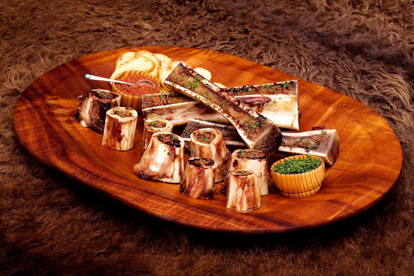 A wooden platter with roasted bone marrow pieces, garnished with herbs. There are small bowls of green herbs and a reddish sauce, along with slices of toasted bread. The platter is set on a textured brown surface.