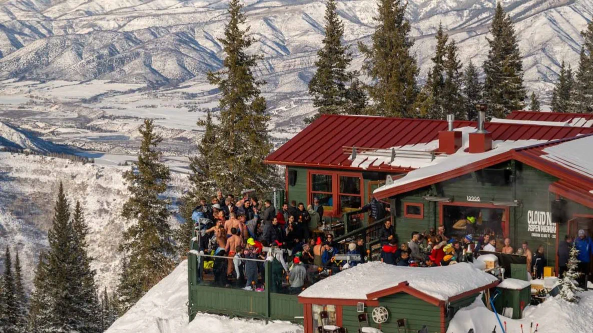 A lively scene at a snowy mountain lodge with a red roof, surrounded by tall evergreen trees. A large group of people is gathered on the outdoor deck, some wearing winter clothing and others in lighter attire, suggesting a festive atmosphere. The background features a stunning view of snow-covered mountains and valleys.