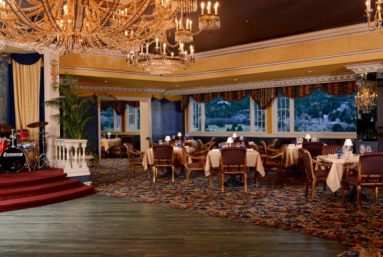 The image shows an elegant restaurant interior with a large, ornate chandelier hanging from the ceiling. The room is decorated with rich colors and patterns, featuring tables set with white tablecloths and blue glassware. There are large windows with views of a scenic landscape outside. A small stage with a drum set is visible on the left side of the image. The overall atmosphere is luxurious and sophisticated.