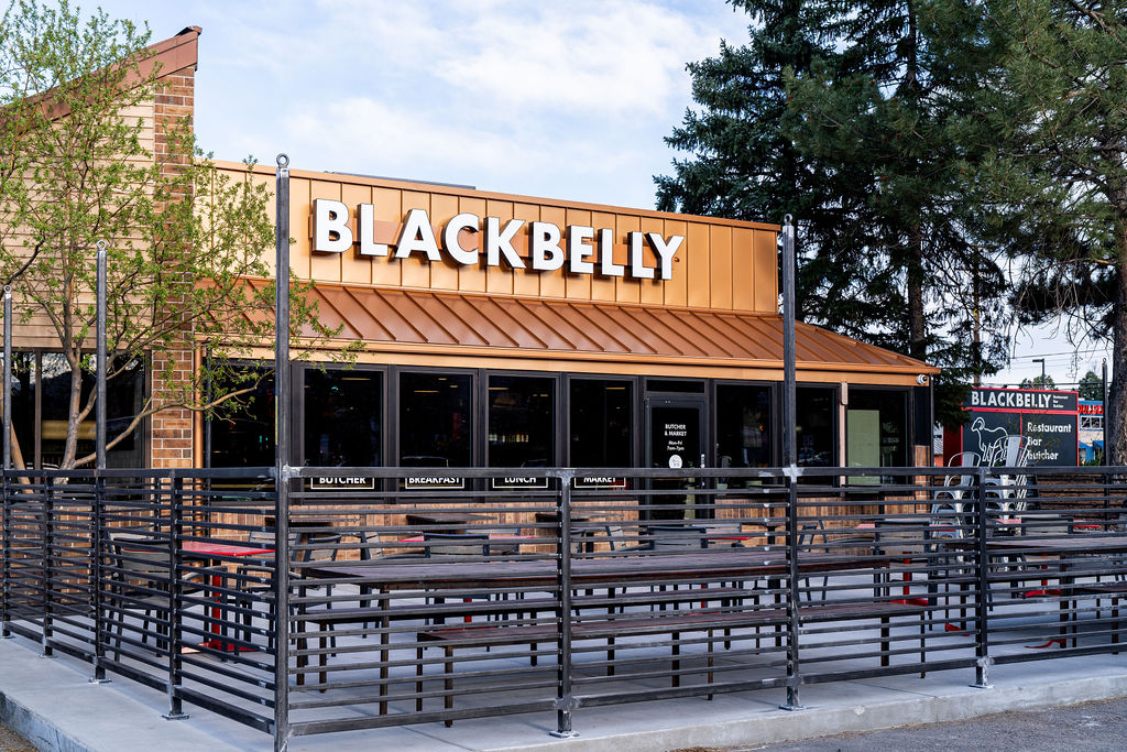 The image shows the exterior of a restaurant named "Blackbelly." The building has a modern design with a brick facade and a metal roof. There is a fenced outdoor seating area with tables and chairs. Trees are visible around the restaurant, and a sign on the right side indicates it is a restaurant, bar, and butcher.