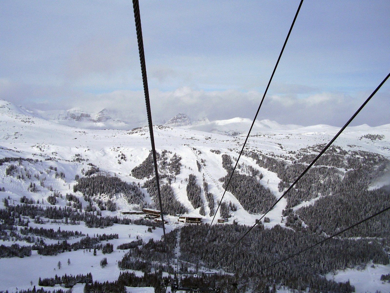 A snowy mountain landscape with ski slopes and a cable car system. The image shows snow-covered trees and mountains under a cloudy sky, with several cables stretching across the scene, indicating a ski lift or gondola.