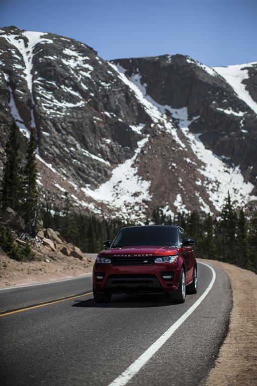 A red Range Rover is driving on a winding mountain road. The background features tall, rocky mountains with patches of snow and scattered evergreen trees under a clear blue sky.