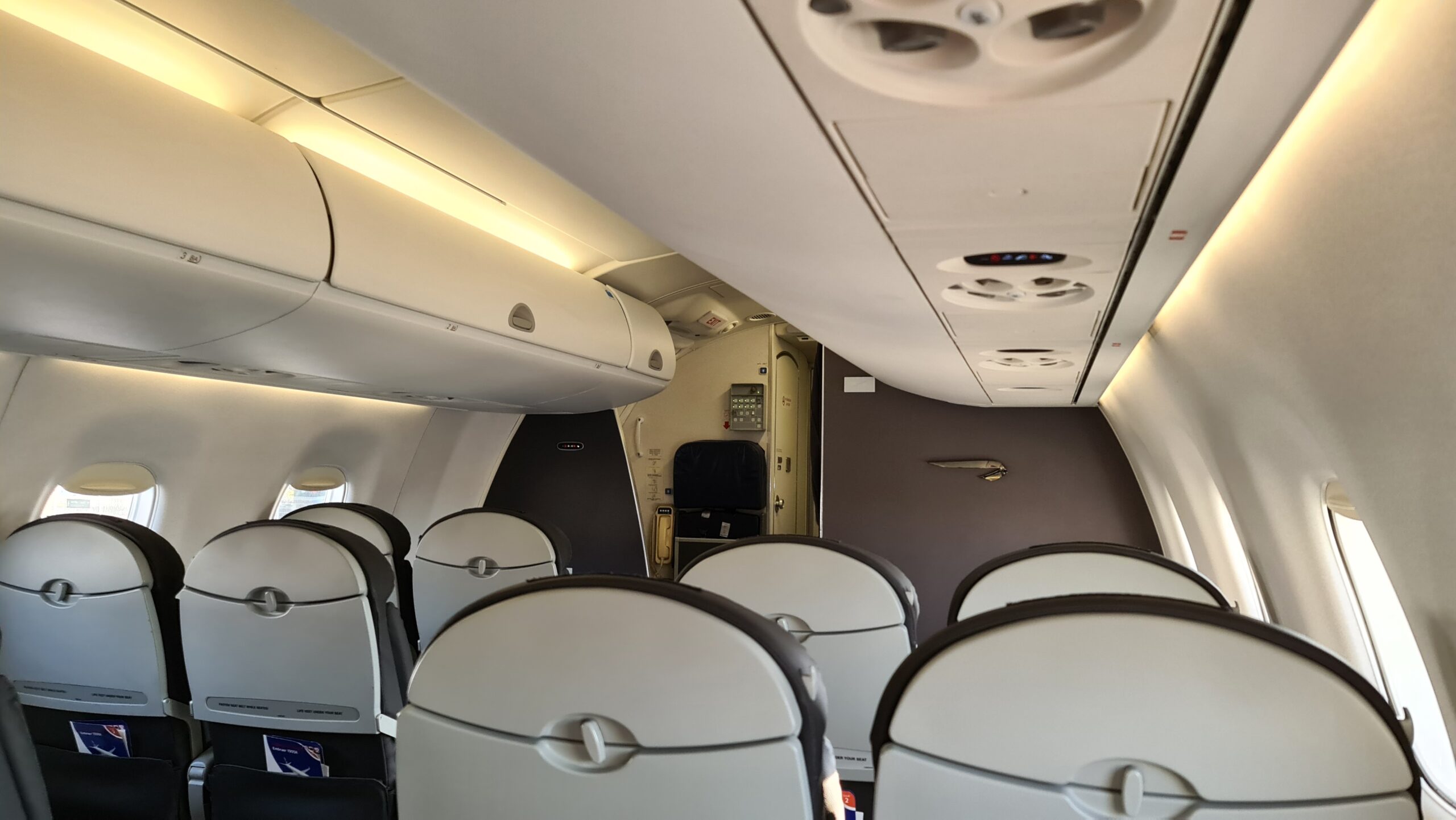 The image shows the interior of an airplane cabin, focusing on the back of several rows of seats. Overhead compartments are closed, and the ceiling has air vents and lights. The cockpit door is visible at the front of the cabin. The cabin is empty, with no passengers present.