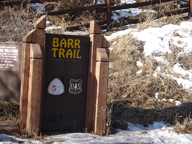 The image shows a wooden sign for "Barr Trail" with two logos below it, one for the U.S. Forest Service and another for the U.S. Department of Agriculture. The sign is surrounded by snow and dry grass, with a wooden fence in the background.