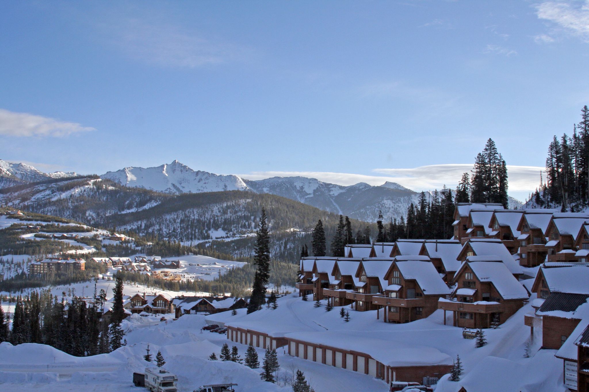 A snowy mountain landscape with a cluster of wooden cabins in the foreground. The cabins have snow-covered roofs and are surrounded by pine trees. In the background, there are more snow-covered mountains under a clear blue sky.