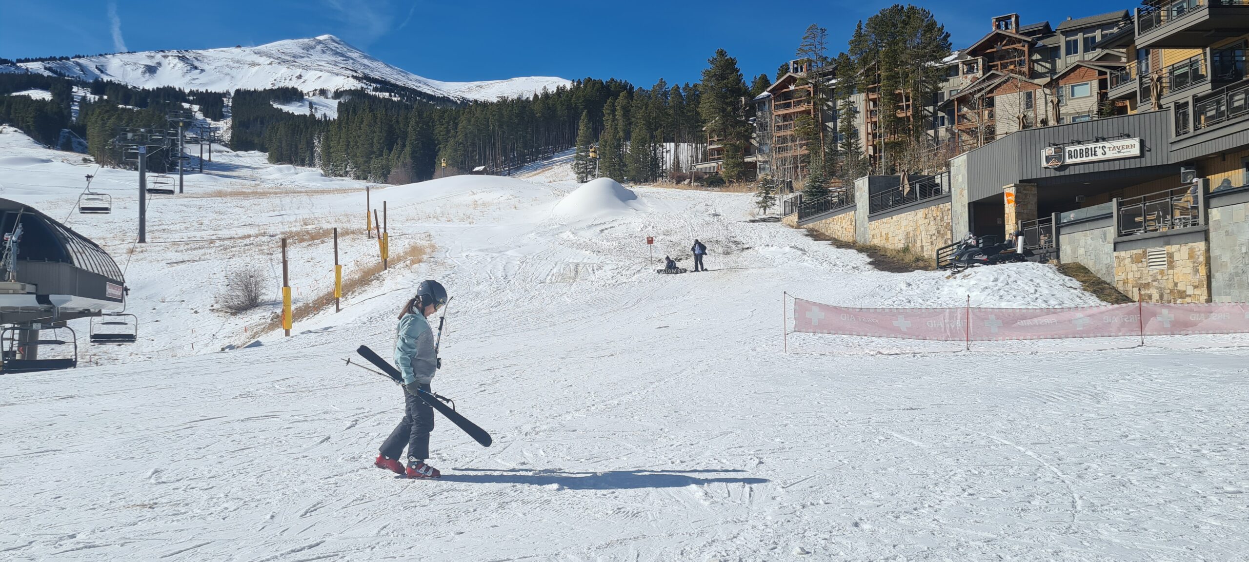 A skier is walking across a snowy slope carrying skis, with a ski lift on the left and a mountain in the background. There are trees and buildings on the right, including a sign for "Robbie's Tavern." The sky is clear and blue.