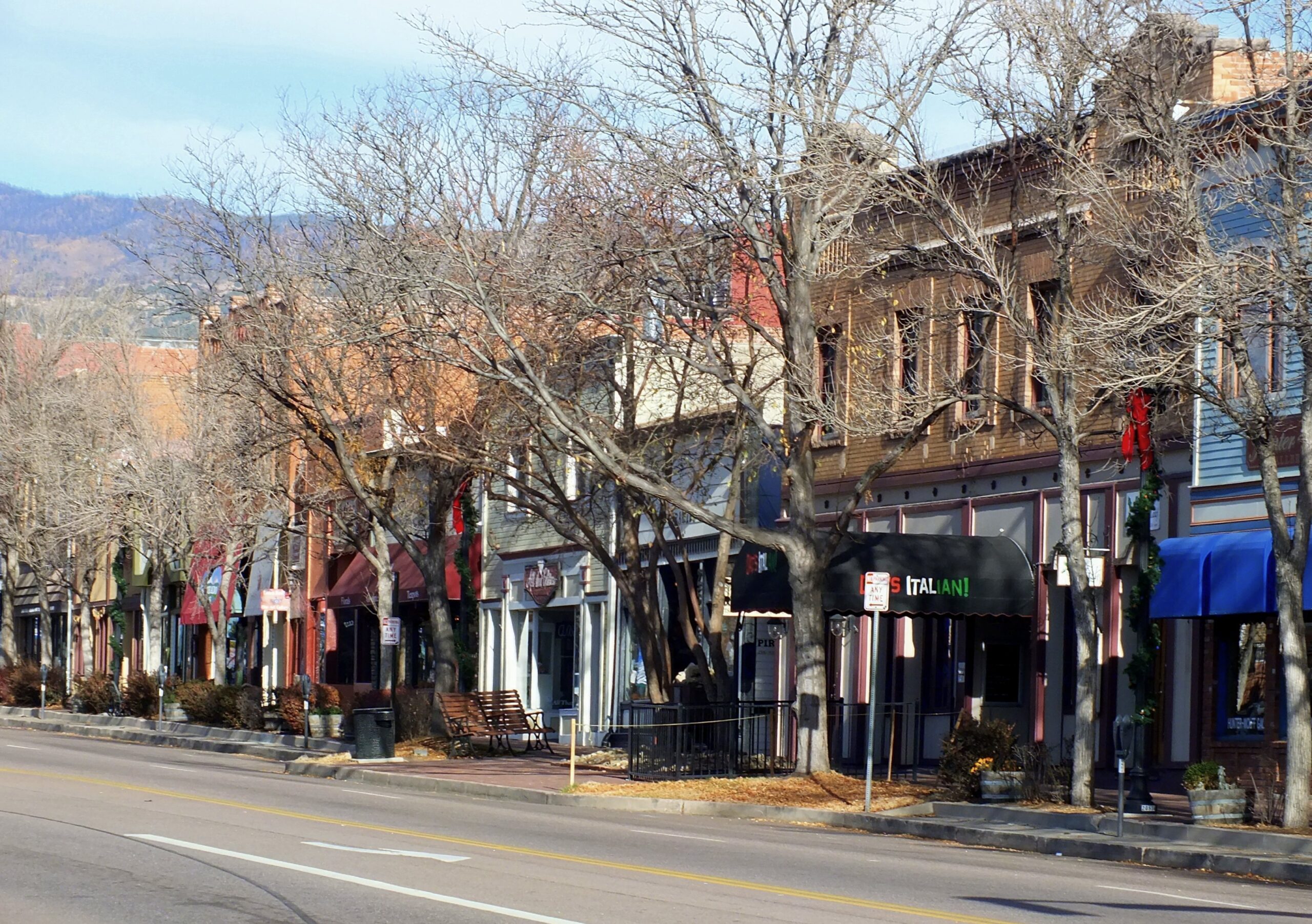 A street view of a small town with a row of colorful storefronts and bare trees lining the sidewalk. The buildings have various awnings and signs, including one for an Italian restaurant. The street is empty, and there are mountains visible in the background.