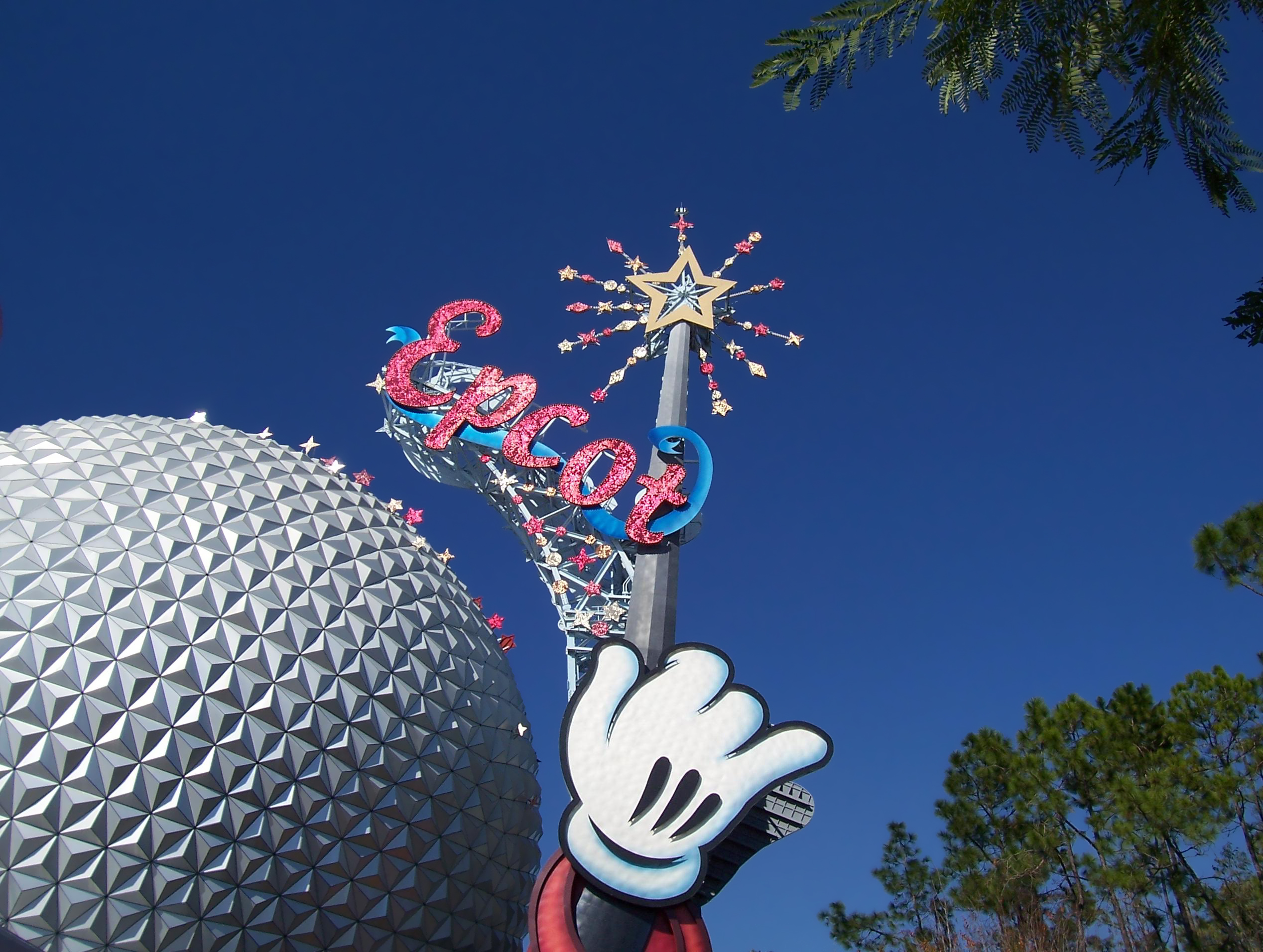 The image shows the iconic Spaceship Earth geodesic sphere at Epcot, part of Walt Disney World Resort. In the foreground, there is a large sign with the word "Epcot" in red, glittery letters, topped with a star and surrounded by smaller stars. A large hand, resembling a cartoon character's glove, is pointing towards the sphere. The sky is clear and blue, with some trees visible on the right side.