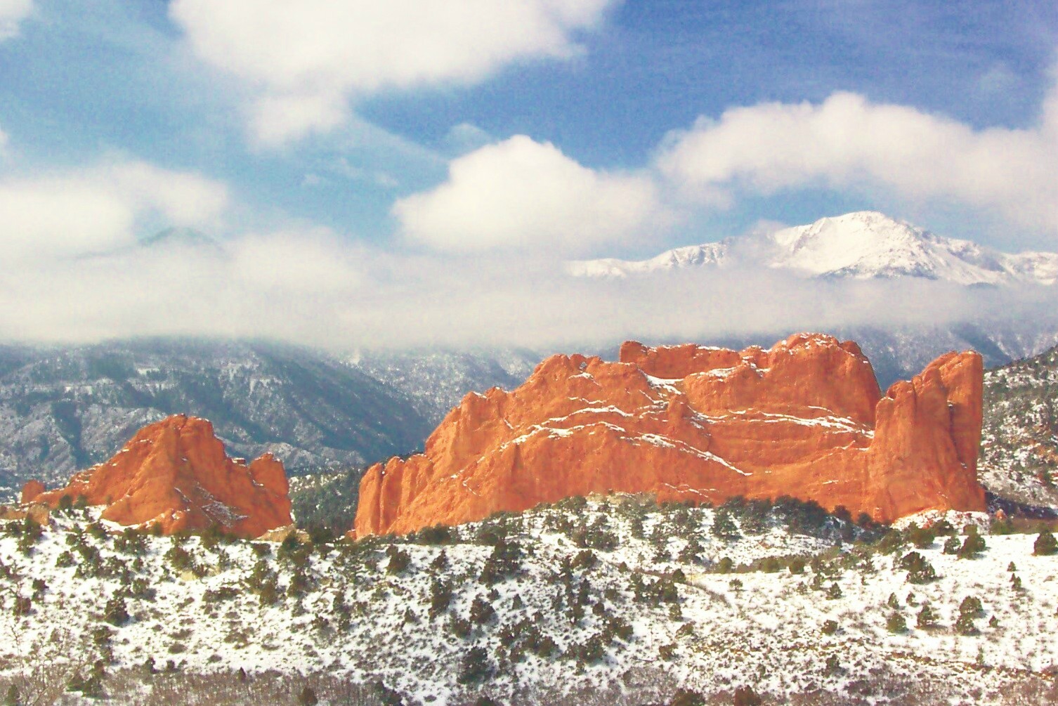 The image shows a scenic landscape featuring red rock formations in the foreground, partially covered with snow. In the background, there are snow-capped mountains under a partly cloudy sky. The scene is likely from a natural park or mountainous region.