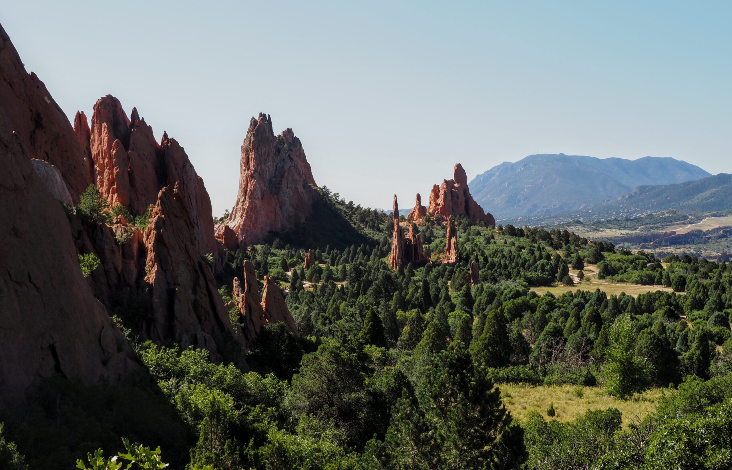 The image shows a scenic landscape featuring tall, jagged red rock formations surrounded by lush green vegetation. In the background, there are distant mountains under a clear blue sky. The scene is likely from a natural park or a similar outdoor setting.