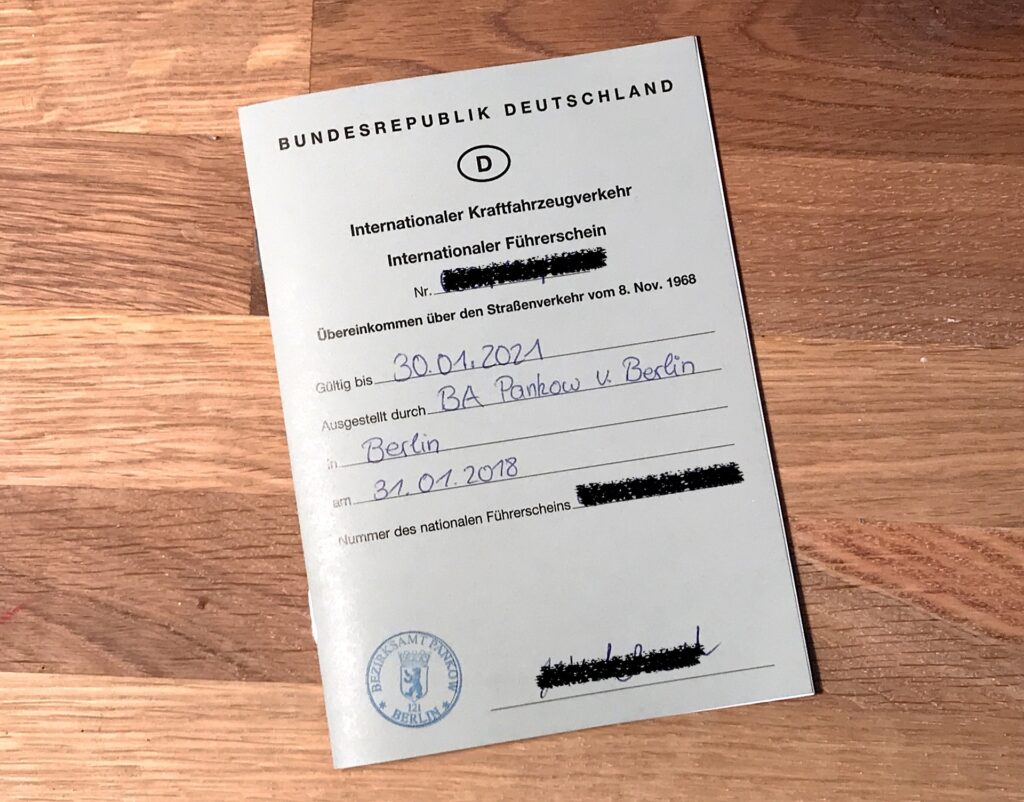 The image shows an International Driving Permit from Germany, titled "BUNDESREPUBLIK DEUTSCHLAND." It includes handwritten details such as the validity date "30.01.2021," the issuing authority "BA Pankow v. Berlin," and the issue date "31.01.2018." Some information is blacked out for privacy. The document is placed on a wooden surface.