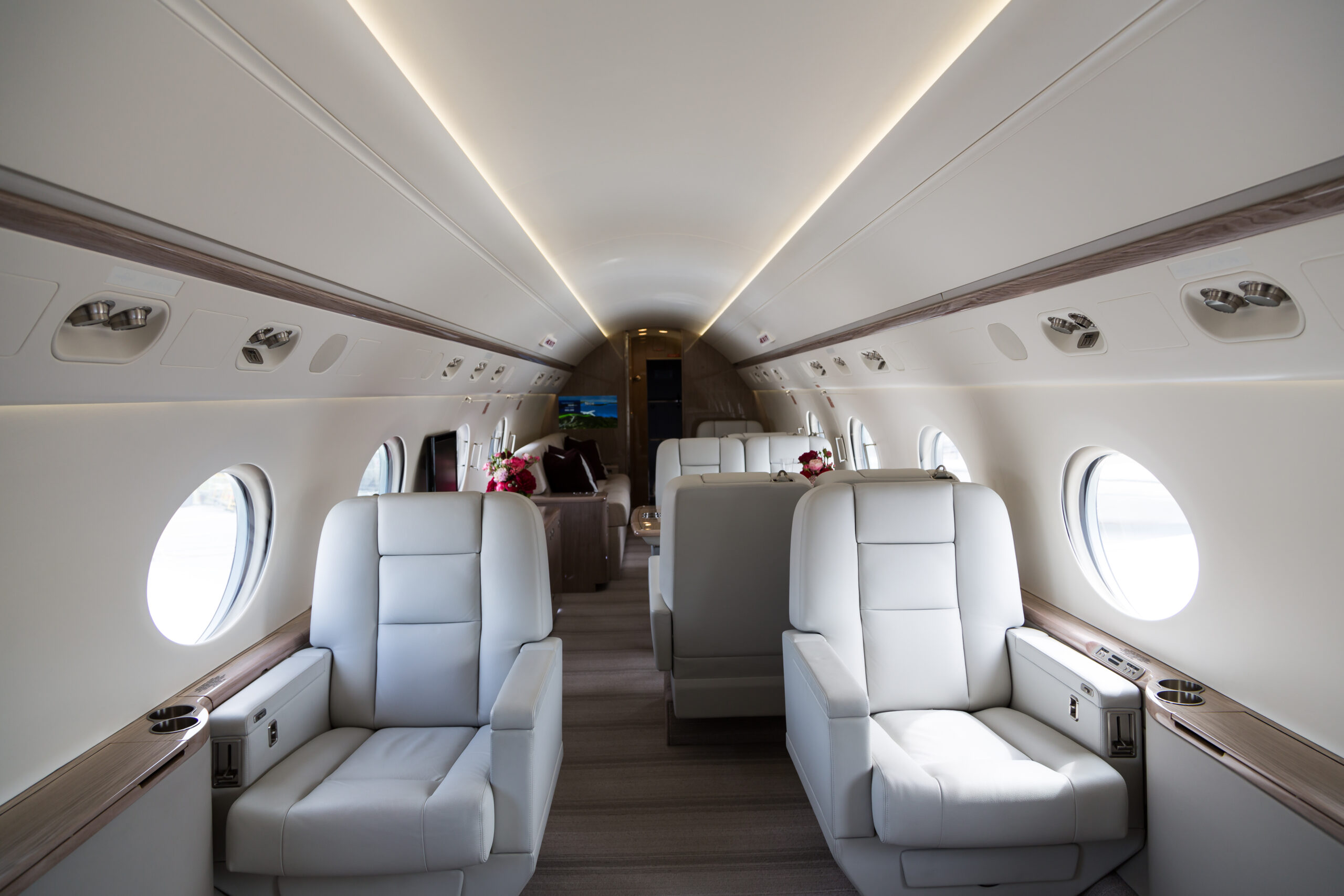 The image shows the interior of a luxurious private jet. It features spacious, plush white leather seats arranged in a comfortable layout. The cabin has large circular windows on both sides, allowing natural light to illuminate the space. There are small tables with flowers and a screen visible in the background, contributing to the elegant and modern design.