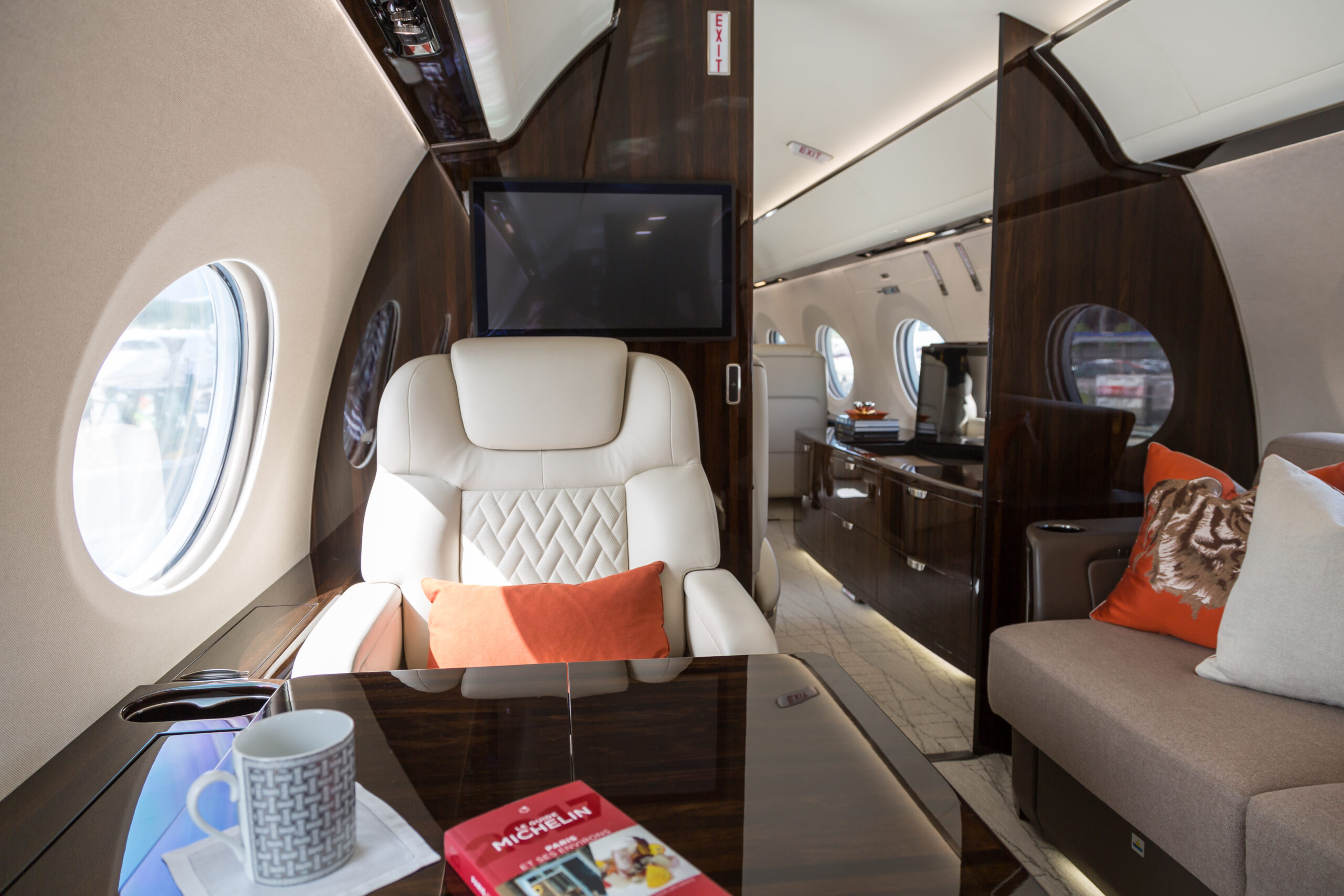 The image shows the interior of a luxurious private jet. It features a plush, cream-colored leather seat with an orange pillow, a wooden table with a patterned mug and a Michelin guidebook, and a sofa with decorative pillows. The cabin has large windows, a flat-screen TV, and elegant wood paneling.