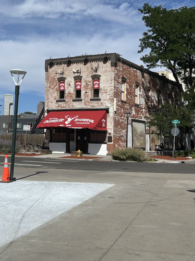 A two-story brick building with a red awning that reads "Buckhorn Exchange Steakhouse" is situated on a street corner. The building has several windows with red signs above them. There are decorative elements, including animal figures, on the facade. A tree is visible on the right, and a traffic cone is on the sidewalk in the foreground. The sky is clear and blue.