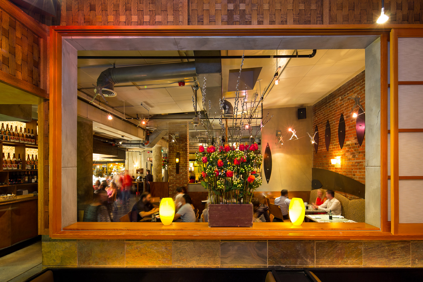 The image shows the interior of a restaurant with a warm, inviting atmosphere. In the foreground, there is a large rectangular vase filled with red roses and decorative branches. The background features a brick wall with modern art pieces and several tables where people are dining. The lighting is soft, with yellow lamps adding to the cozy ambiance.