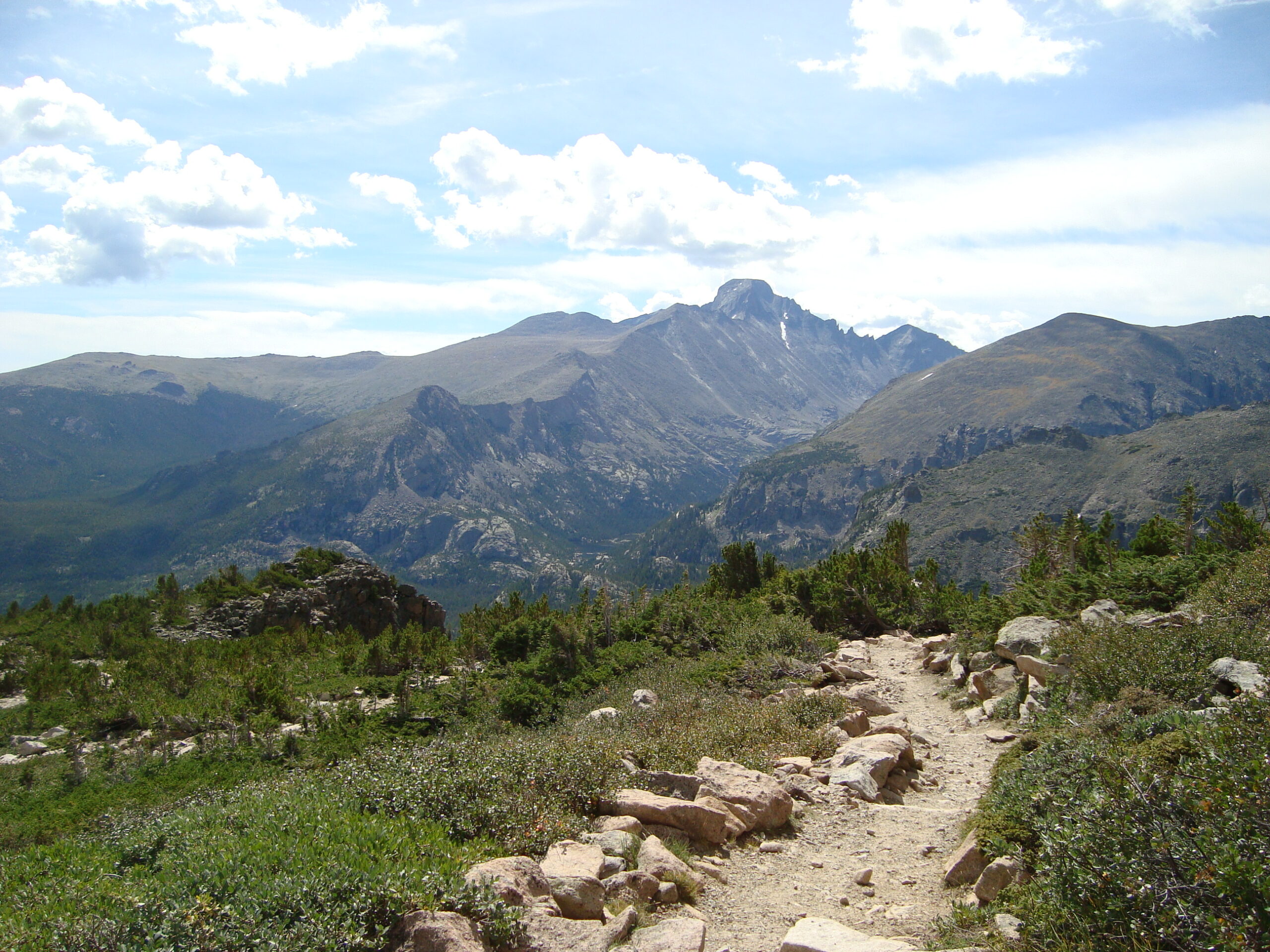 A scenic mountain landscape with a rocky trail in the foreground, surrounded by green vegetation. In the background, there are rugged mountain peaks under a partly cloudy sky.