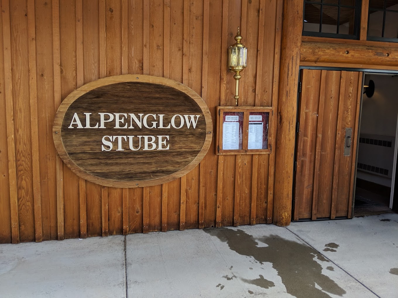 The image shows a wooden exterior wall with a large oval sign that reads "ALPENGLOW STUBE." Next to the sign is a brass lantern and two framed menu displays. The entrance to the building is partially visible with a sliding wooden door slightly open. There are wet patches on the concrete ground in front of the entrance.