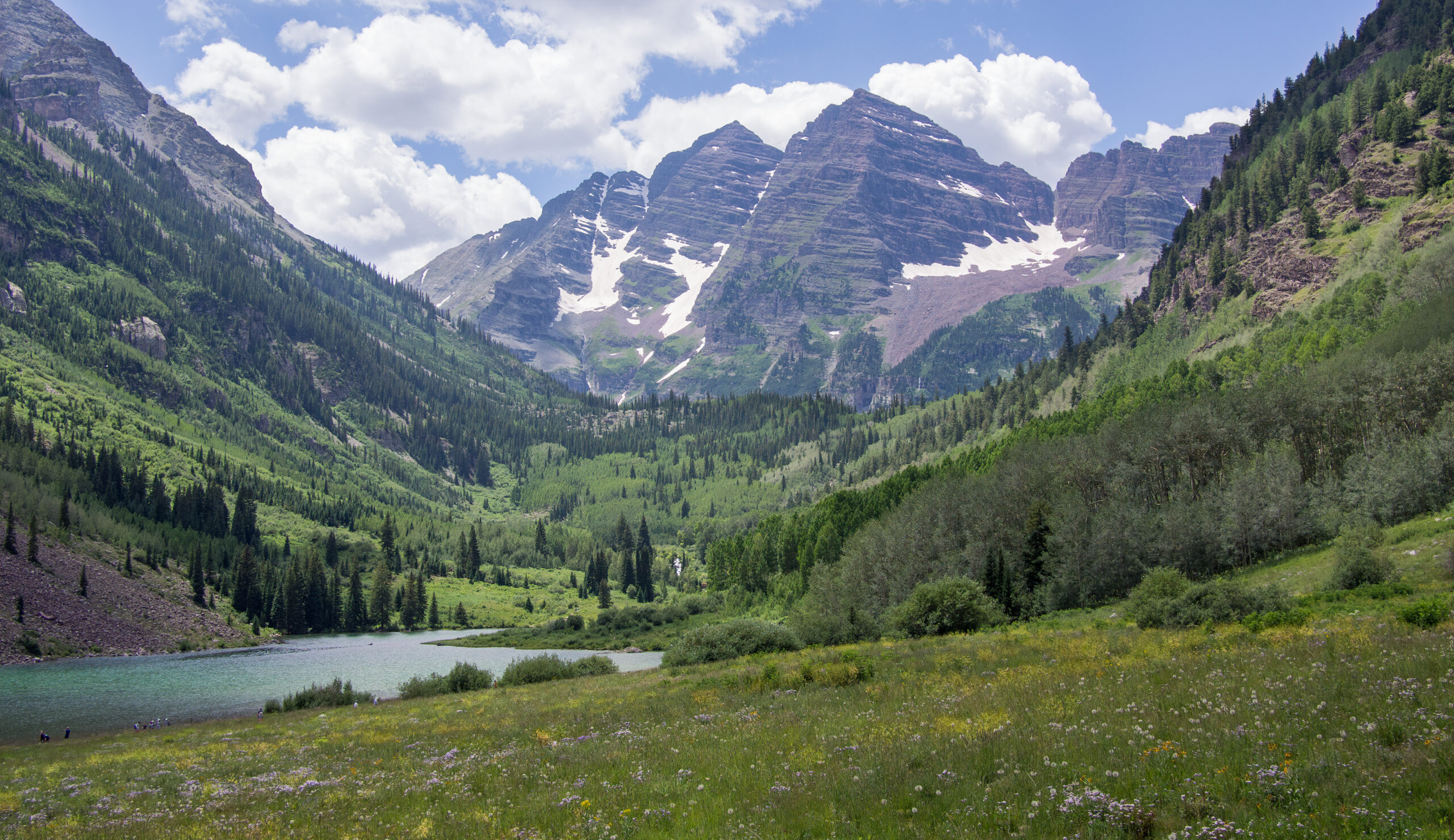 A scenic landscape featuring a lush green valley with a small lake in the foreground. The valley is surrounded by forested hills and towering mountains in the background, partially covered with patches of snow. The sky is blue with scattered white clouds.