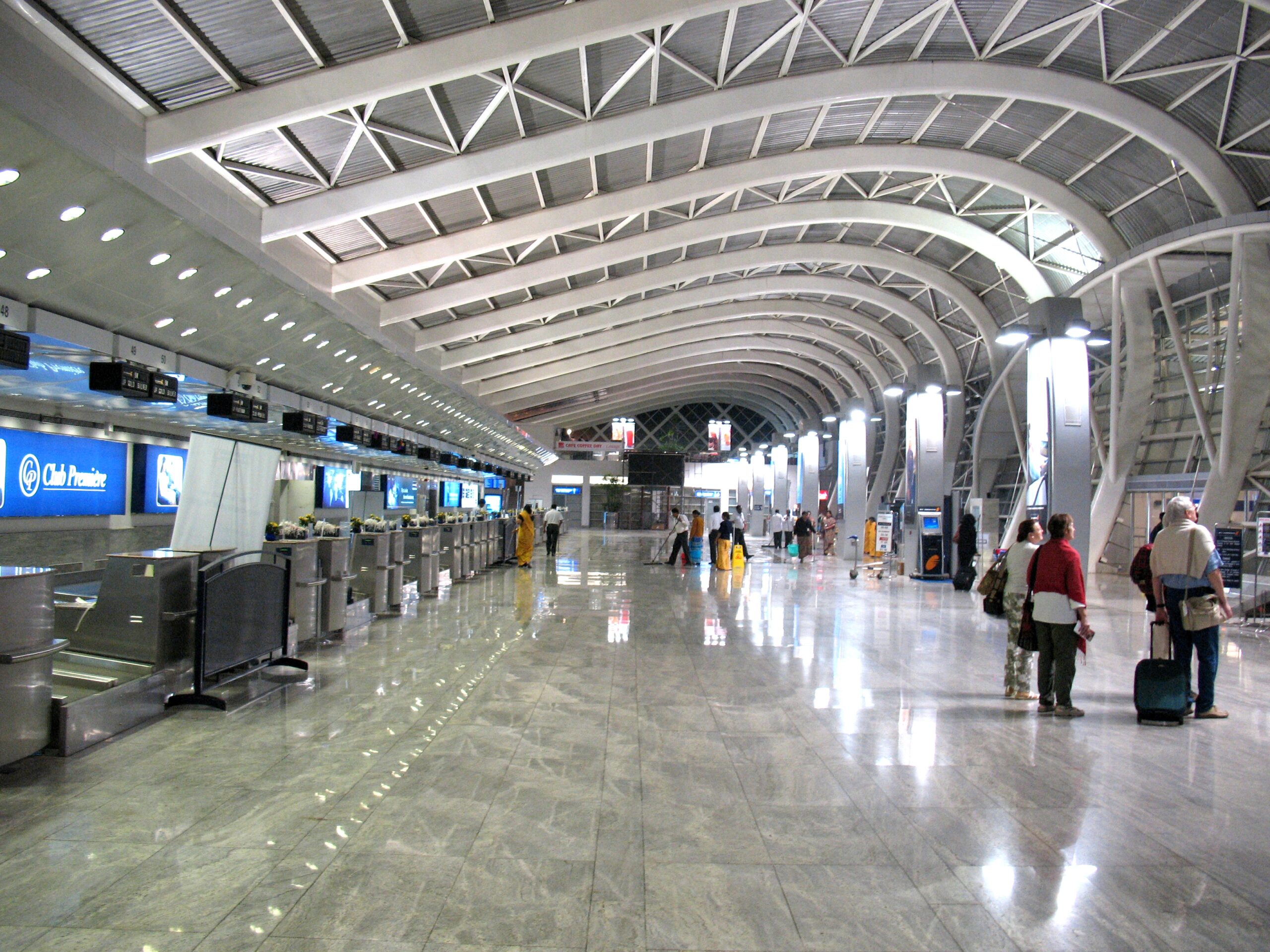 The image shows the interior of a modern airport terminal. The terminal features a high, curved ceiling with exposed beams and a spacious, polished floor. On the left side, there are check-in counters with digital screens above them. Several people are walking or standing with luggage, and the area is well-lit with overhead lights. The architecture is sleek and contemporary.