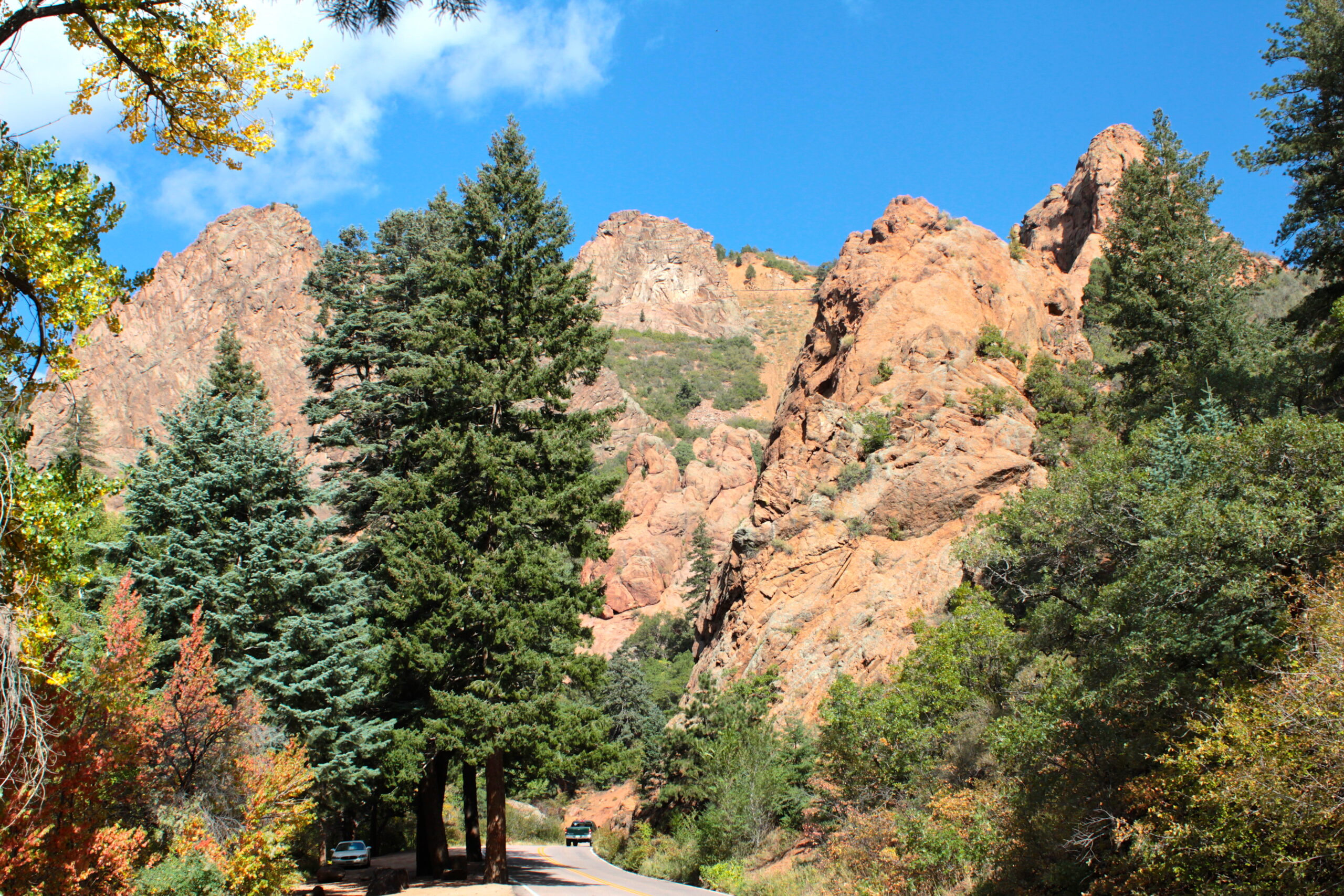 A scenic view of a mountainous landscape with large rock formations and evergreen trees. A winding road runs through the scene, with a car visible on it. The sky is clear and blue, and there are some autumn-colored leaves on the trees.