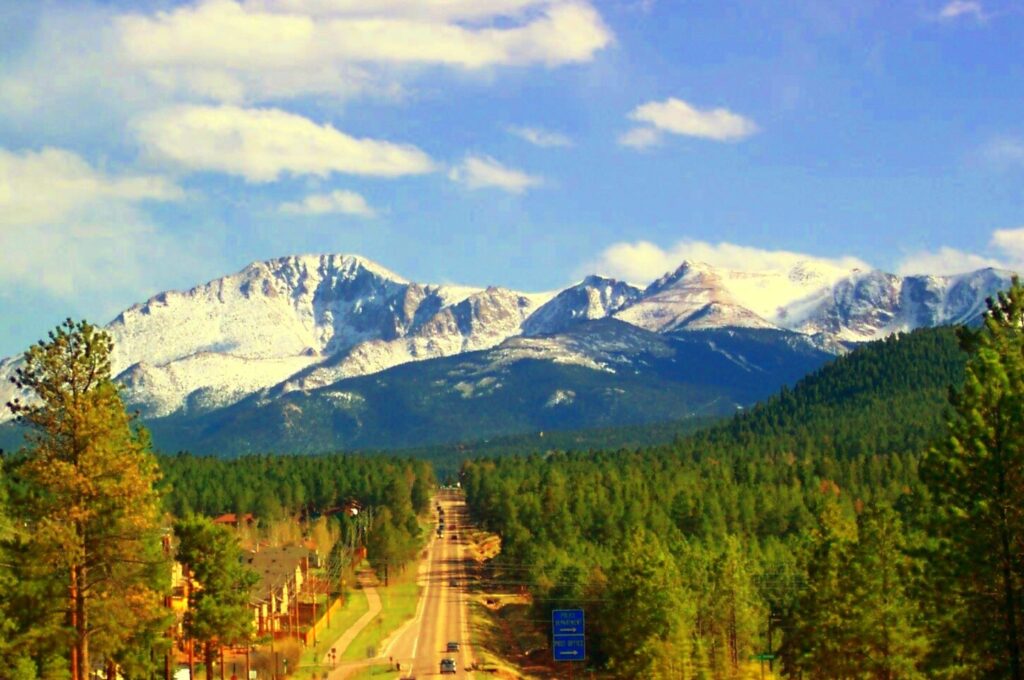 A scenic view of a long road leading towards a range of snow-capped mountains under a blue sky with scattered clouds. The foreground is filled with dense green pine trees, and there are a few buildings visible on the left side of the road.