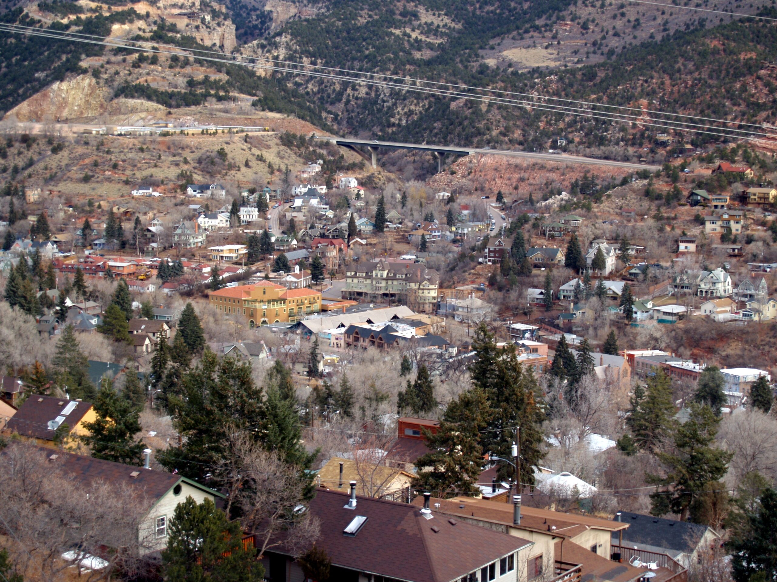 A small town nestled in a mountainous area, with numerous houses and buildings surrounded by trees. The landscape is hilly, with a mix of evergreen and deciduous trees. A bridge and road are visible in the background, crossing over a valley. The scene has a rustic and serene atmosphere.