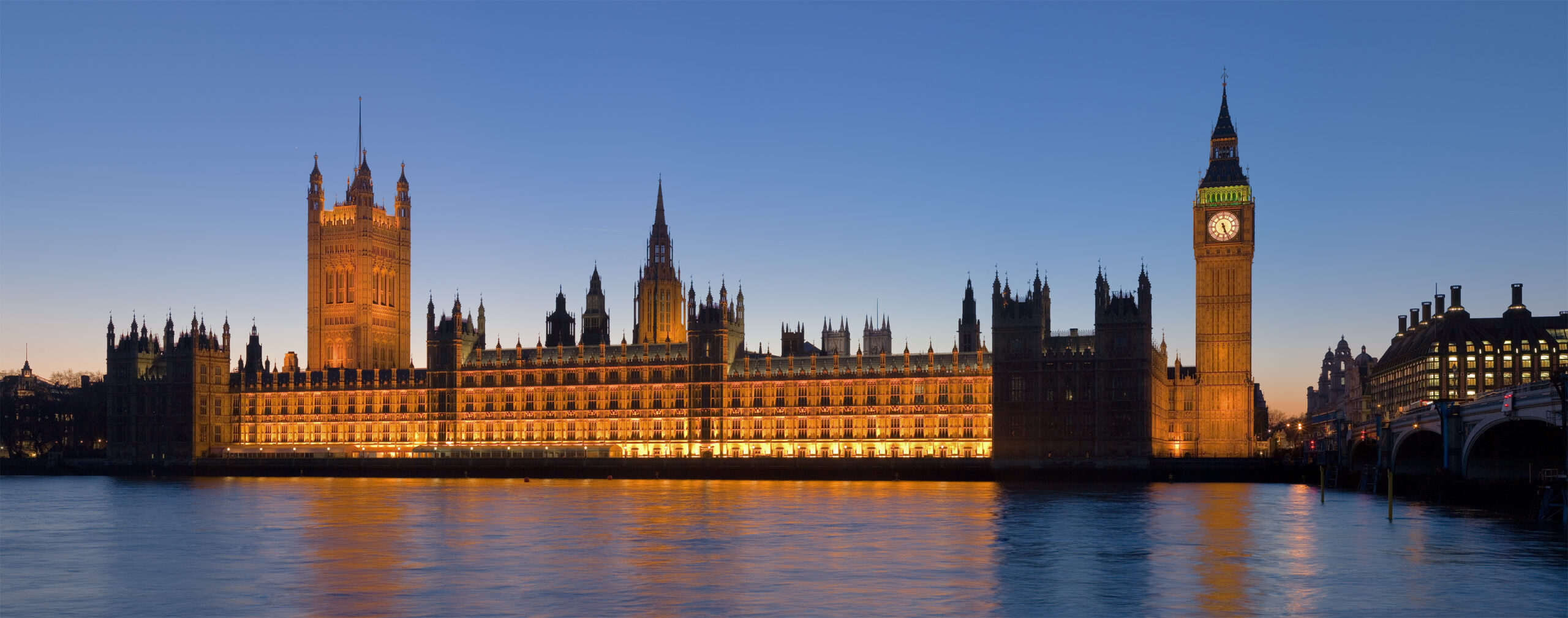 The image shows the Palace of Westminster in London, illuminated at dusk. The iconic clock tower, commonly known as Big Ben, is prominently visible. The building is reflected in the River Thames in the foreground, and the sky is a clear blue as the sun sets.