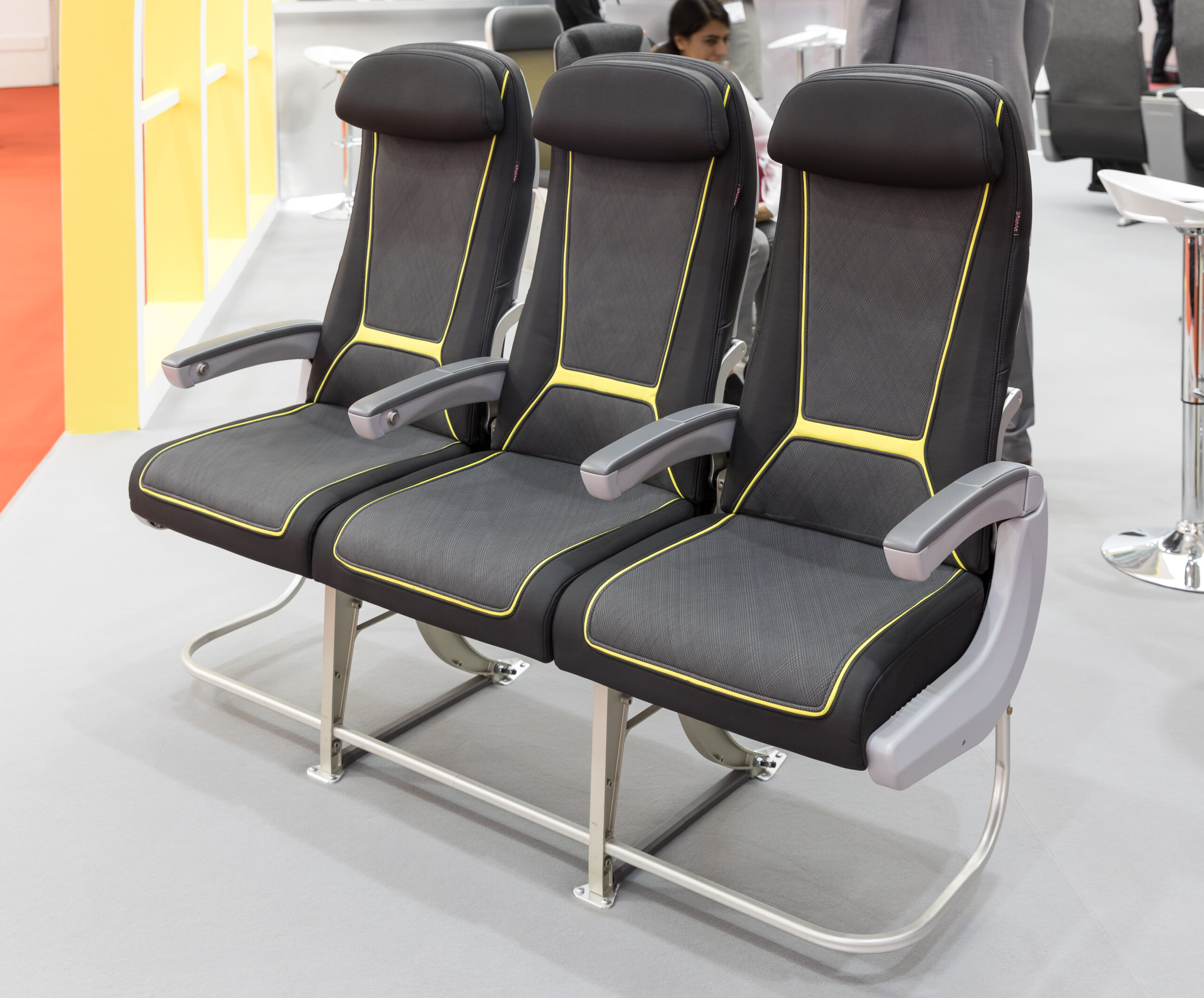 The image shows a row of three modern, black and gray seats with yellow accents, likely designed for an airplane or a similar setting. The seats have armrests and headrests, and they are displayed on a light gray floor with a red carpet and yellow wall in the background.