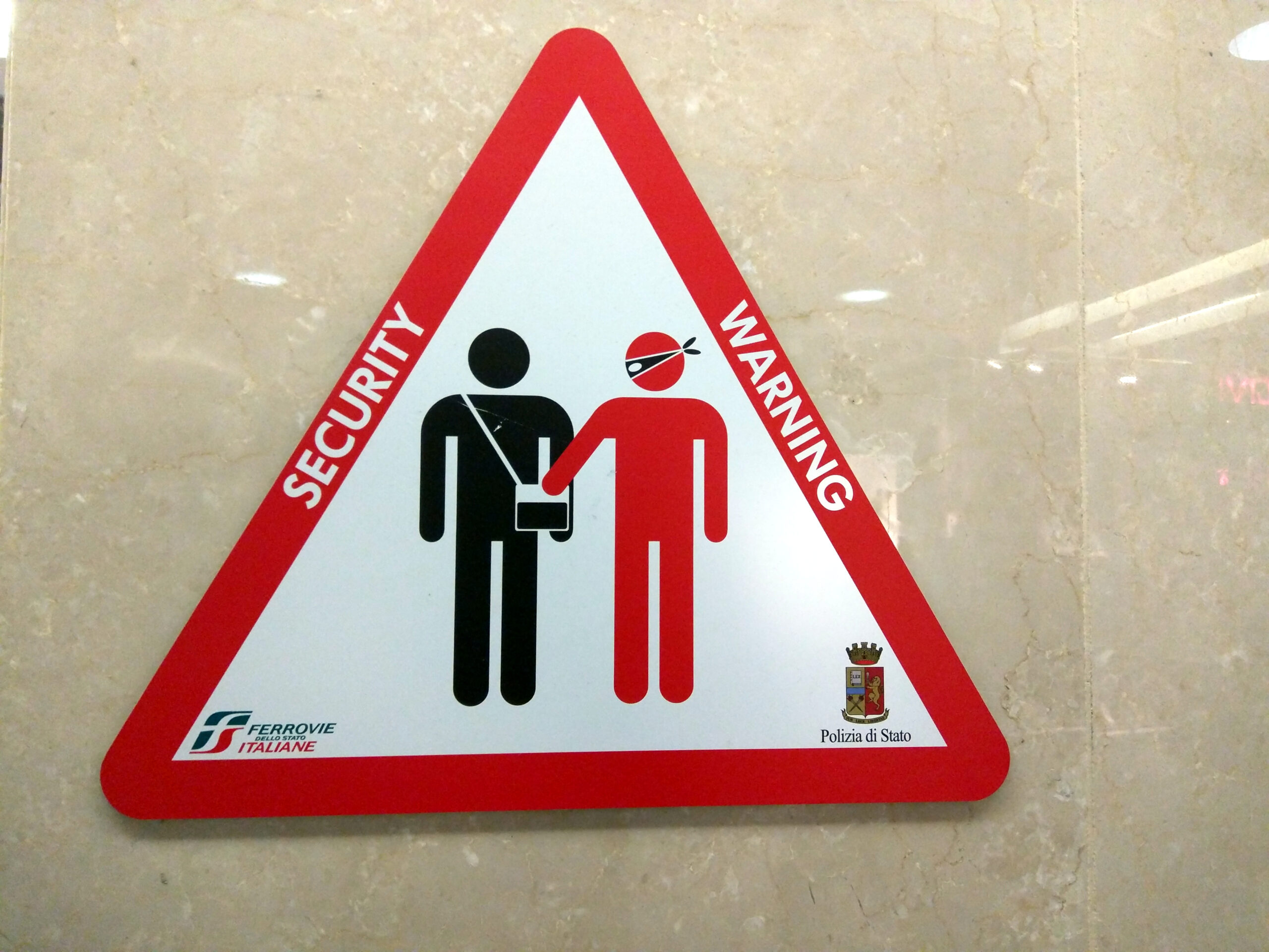 The image shows a triangular security warning sign with a red border. Inside, there are two stylized figures: a black figure with a shoulder bag and a red figure wearing a mask, reaching into the black figure's bag. The text "SECURITY WARNING" is written along the border. Logos for "Ferrovie Italiane" and "Polizia di Stato" are at the bottom.