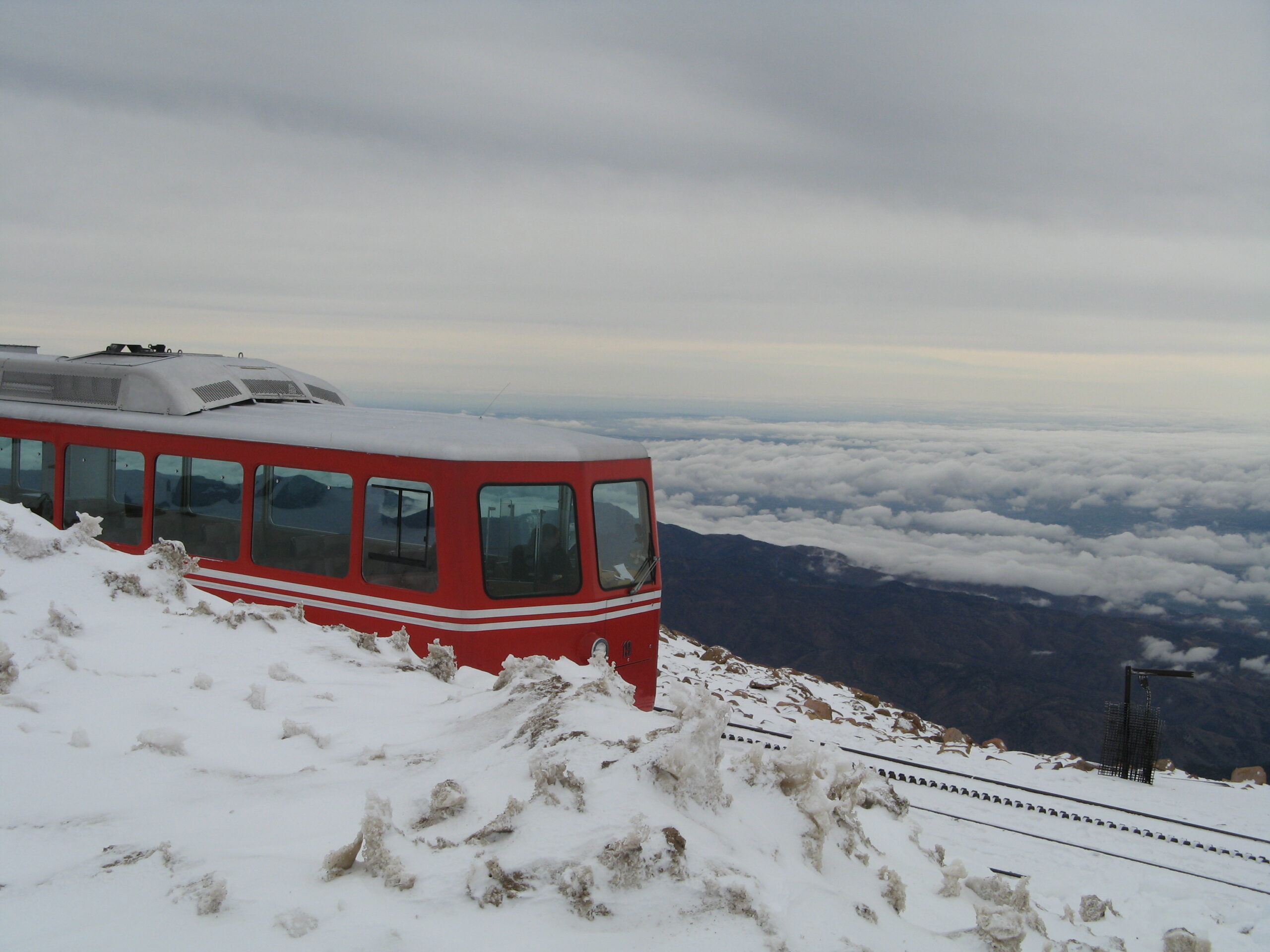 A red train is traveling on a snowy mountain track, with a view of clouds and distant mountains in the background. The sky is overcast, and the scene is serene and elevated.