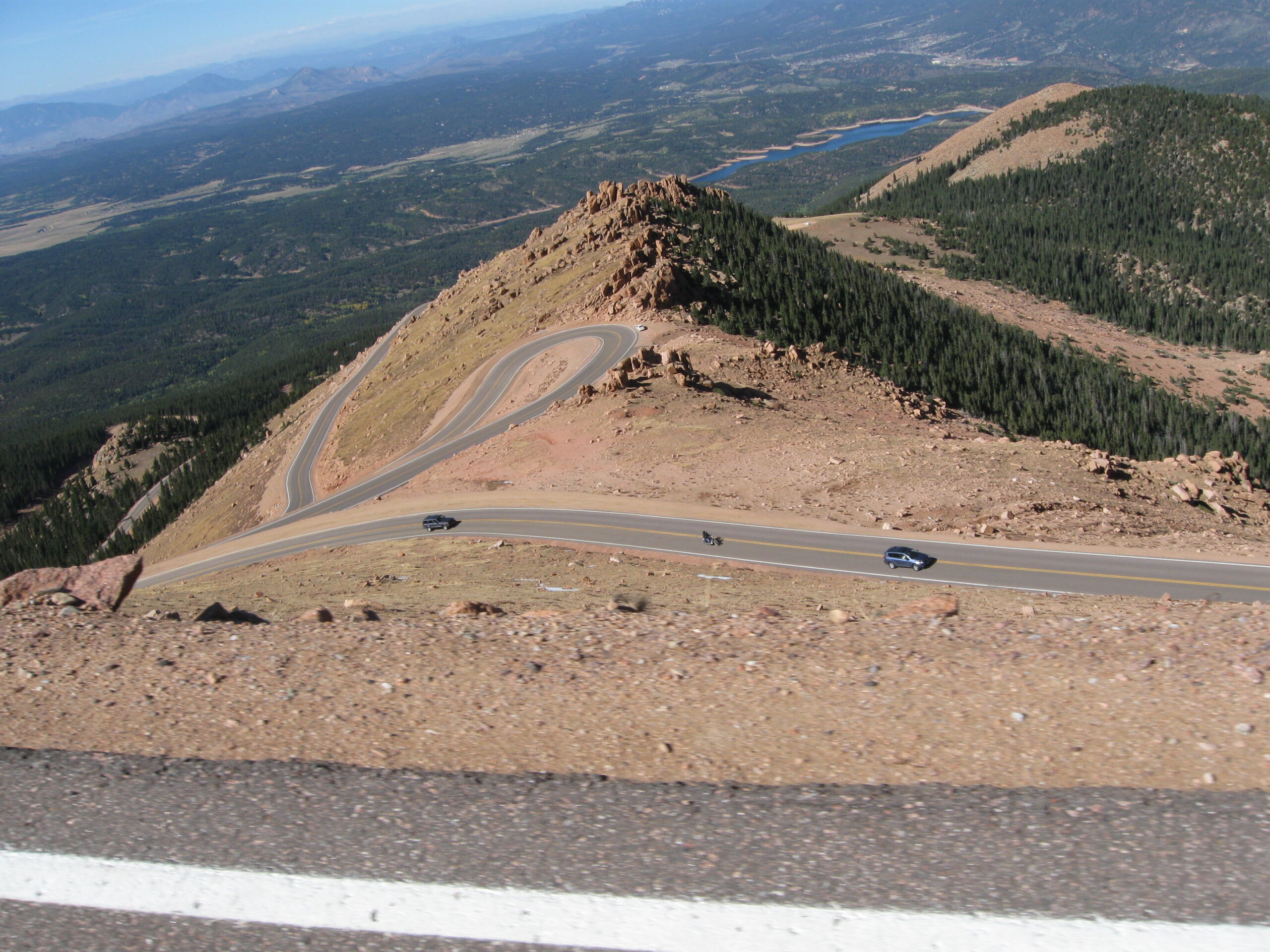 A winding mountain road with several sharp curves is visible, surrounded by rocky terrain and sparse vegetation. Two cars and a motorcycle are traveling along the road. In the background, there are dense forests and a distant view of mountains and a body of water under a clear blue sky.