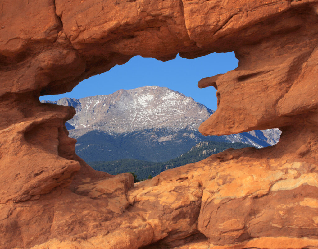 A mountain with a snow-dusted peak is visible through a natural rock window. The rock formation in the foreground is reddish-brown, contrasting with the clear blue sky and the mountain in the background.