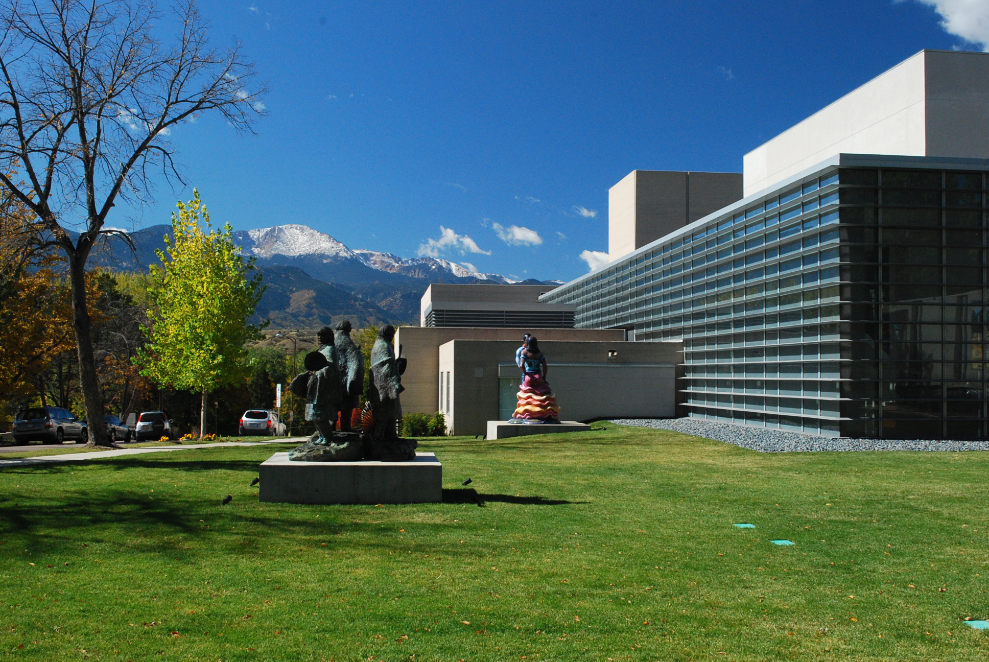 The image shows a modern building with large glass windows on the right side, set against a backdrop of snow-capped mountains under a clear blue sky. In the foreground, there is a grassy area with two sculptures. One sculpture depicts a group of figures, and the other is a colorful abstract form. Trees with autumn foliage and parked cars are visible on the left.