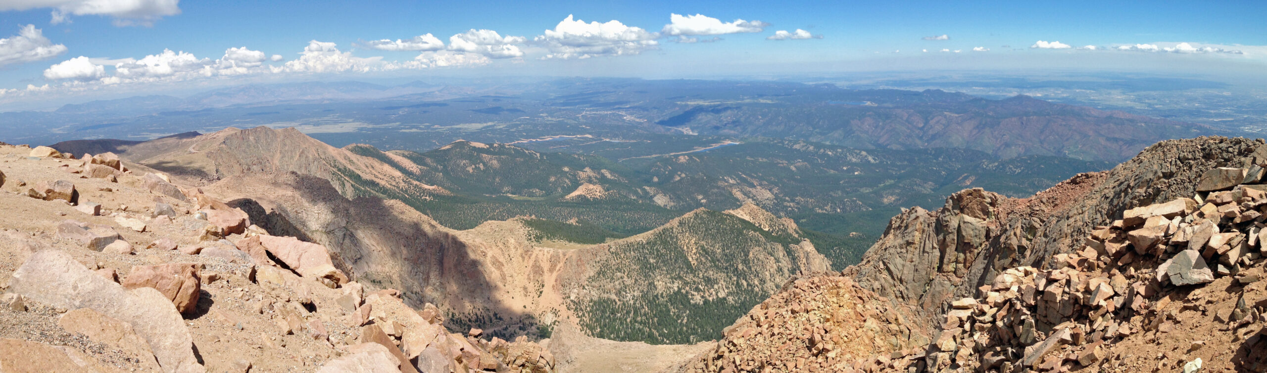 A panoramic view from a rocky mountain summit, showing a vast landscape of rolling hills and valleys covered with green forests. The sky is clear with scattered white clouds, and the horizon stretches far into the distance. The foreground features rugged, rocky terrain.