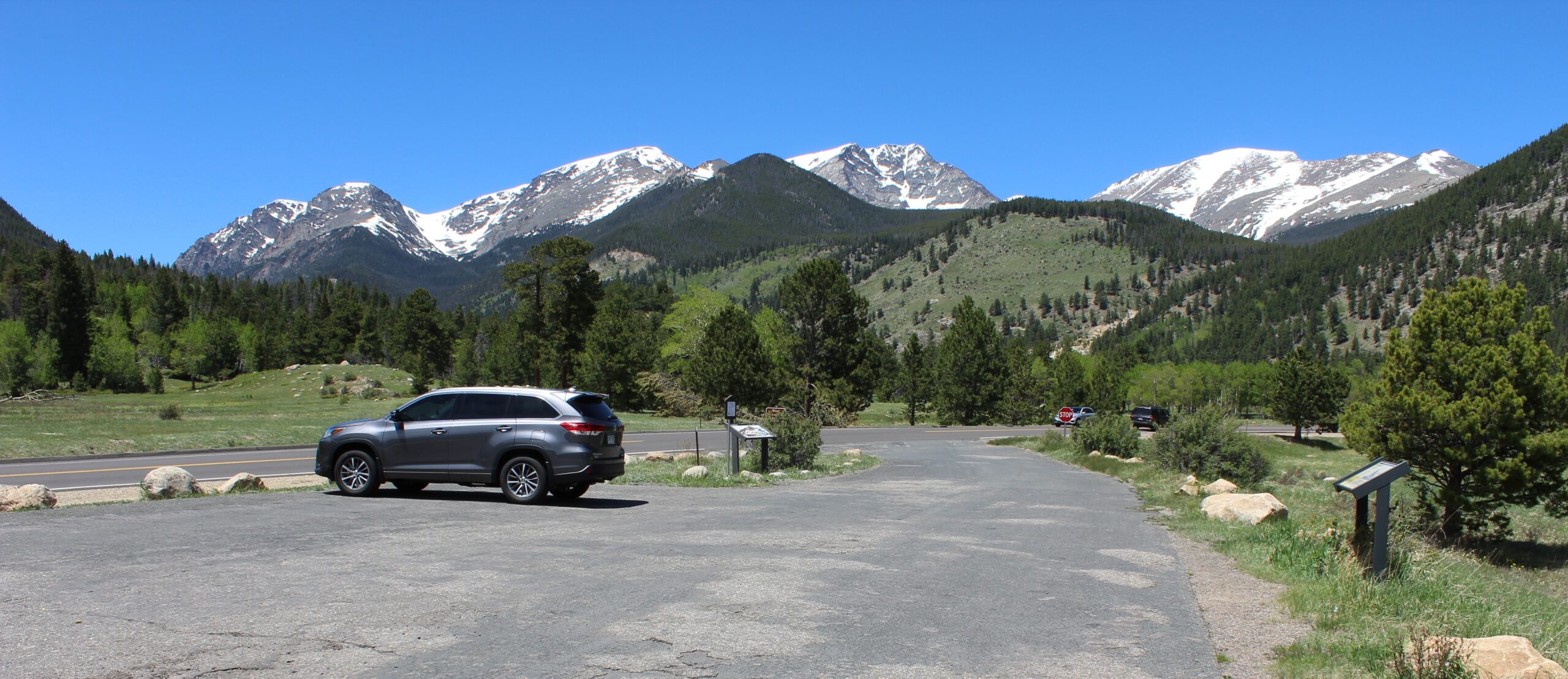 A scenic view of a mountainous landscape with snow-capped peaks in the background under a clear blue sky. In the foreground, there is a parking area with a silver SUV parked on the left. The area is surrounded by lush green trees and grass, with a road and informational signs visible.