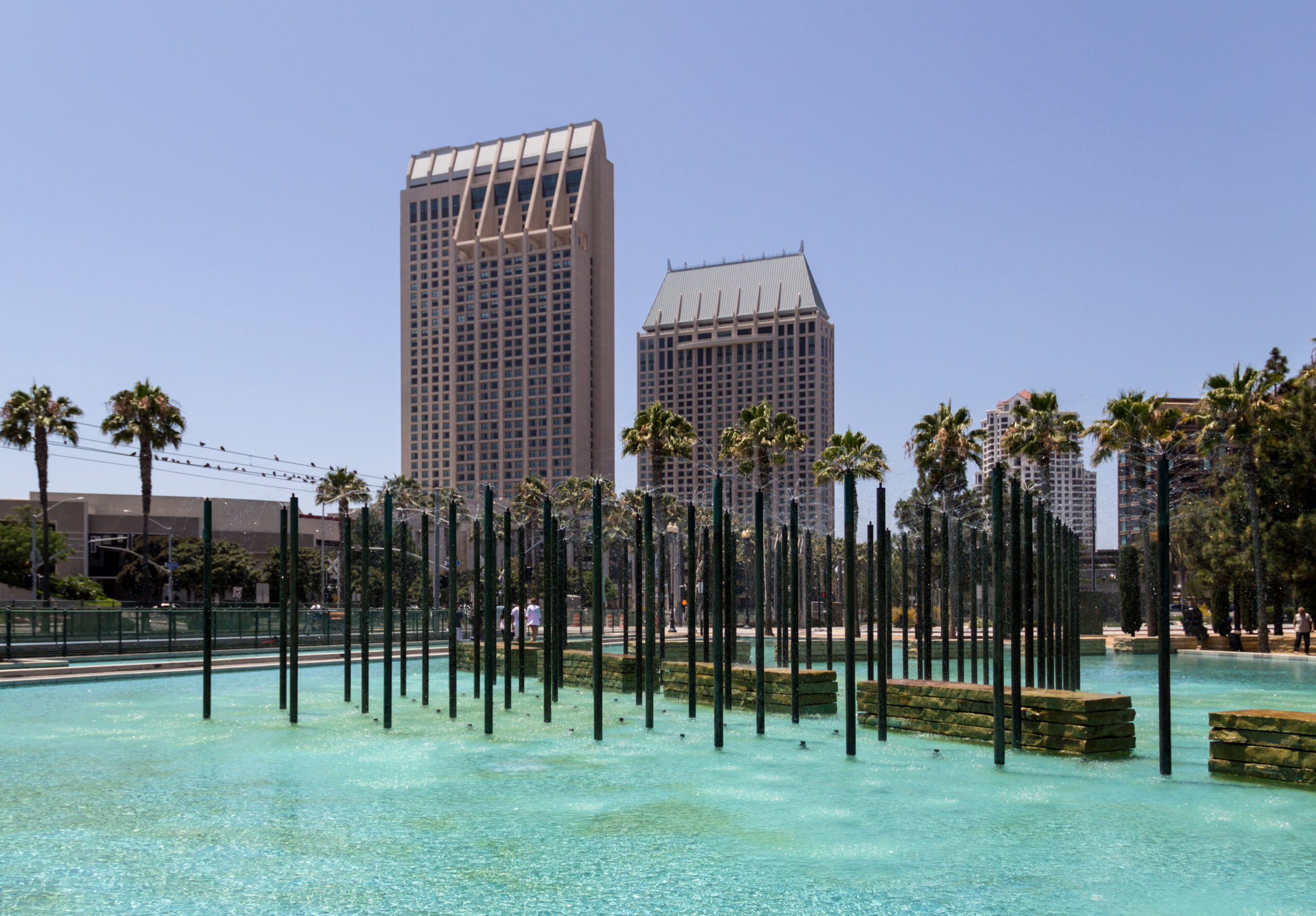 The image shows a cityscape with two tall buildings in the background. In the foreground, there is a large pool of clear blue water with vertical metal poles and water fountains. Palm trees line the area, and the sky is clear and blue.