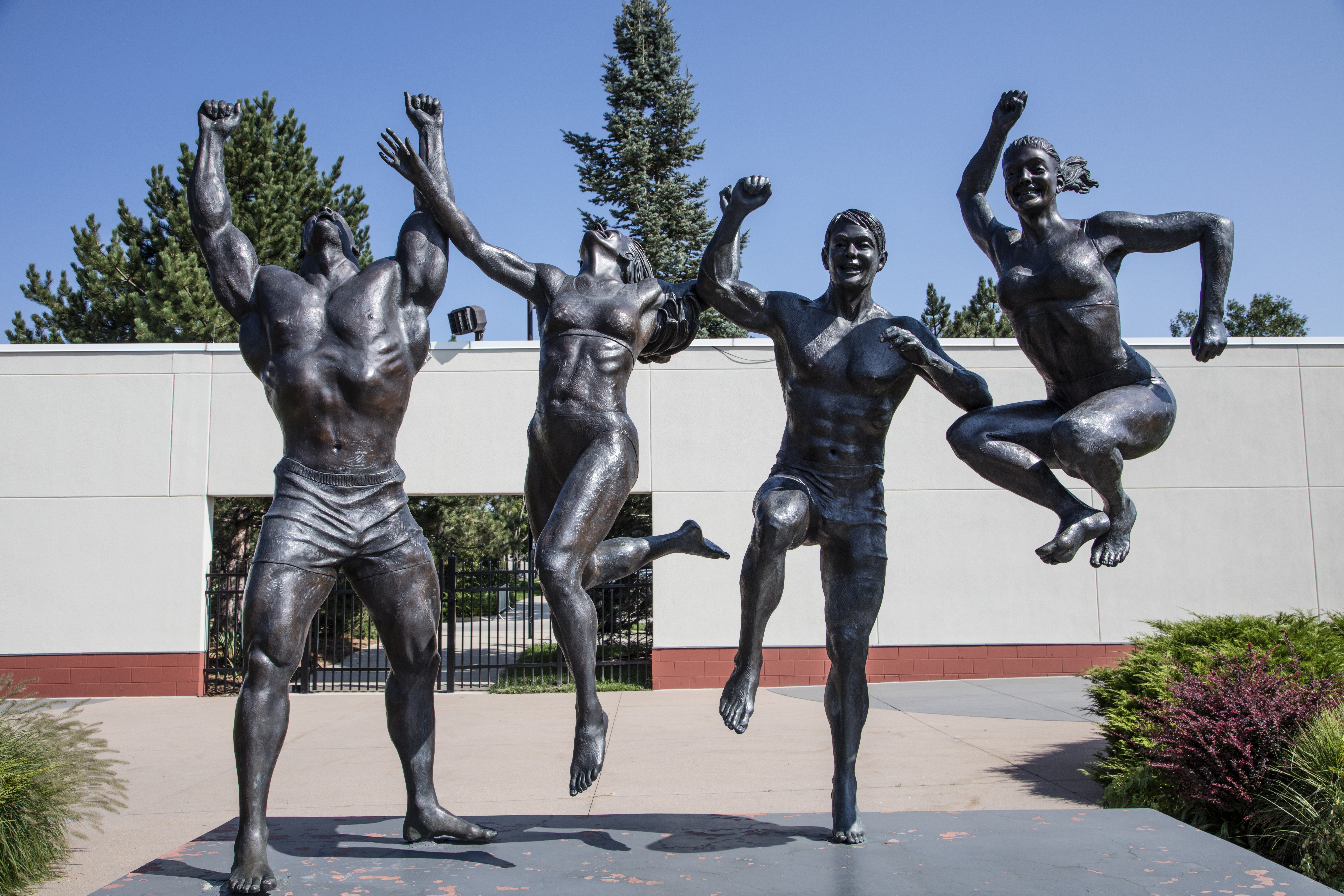 The image shows a bronze sculpture of four athletic figures in dynamic, joyful poses. They appear to be leaping or dancing with their arms raised. The sculpture is set outdoors, with trees and a building in the background.