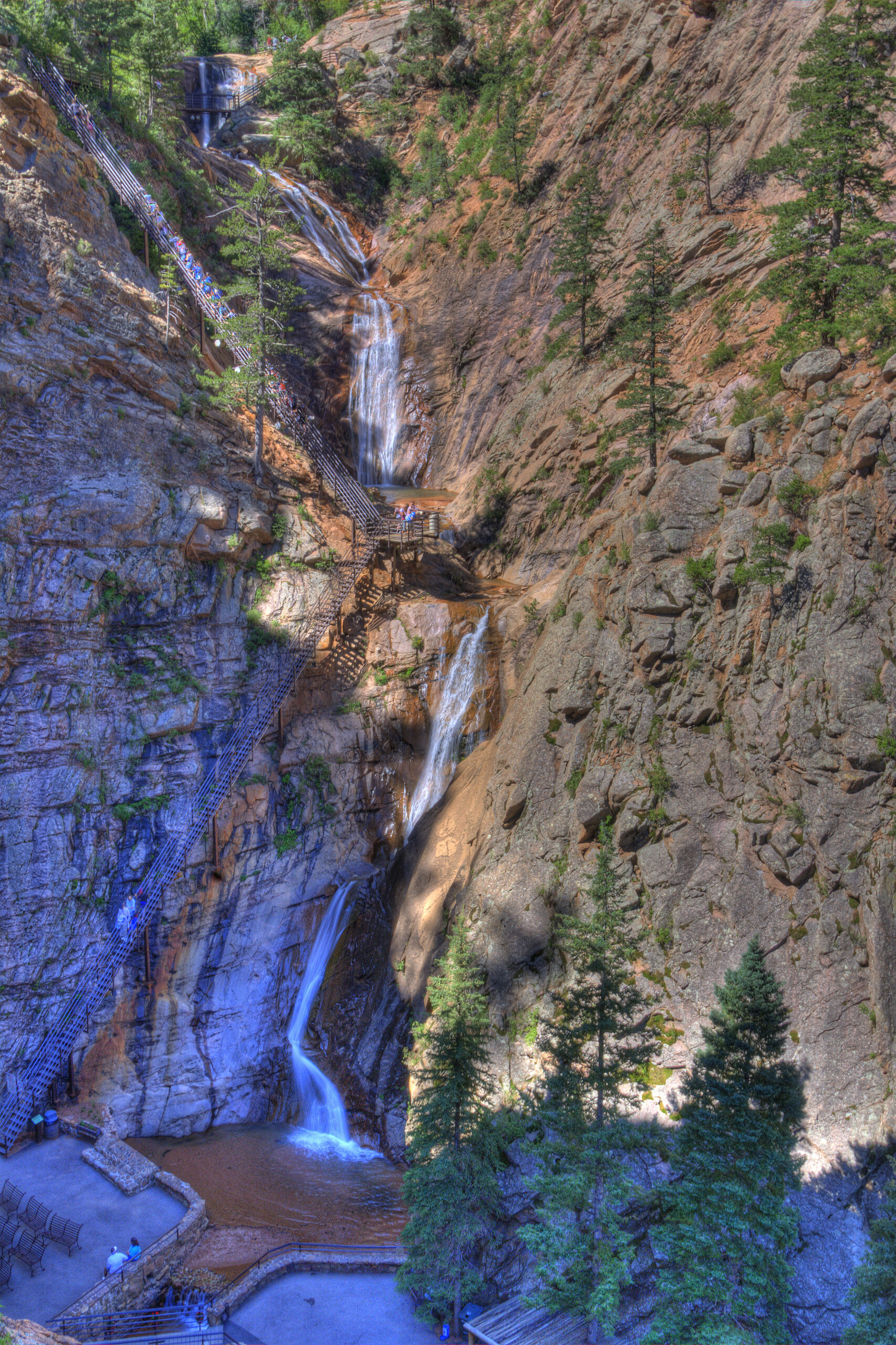 The image shows a tall, multi-tiered waterfall cascading down a rocky cliff surrounded by trees. There are staircases and viewing platforms along the cliff, allowing people to observe the waterfall. At the base, there is a small pool of water and a viewing area with benches. The scene is set in a natural, forested environment.