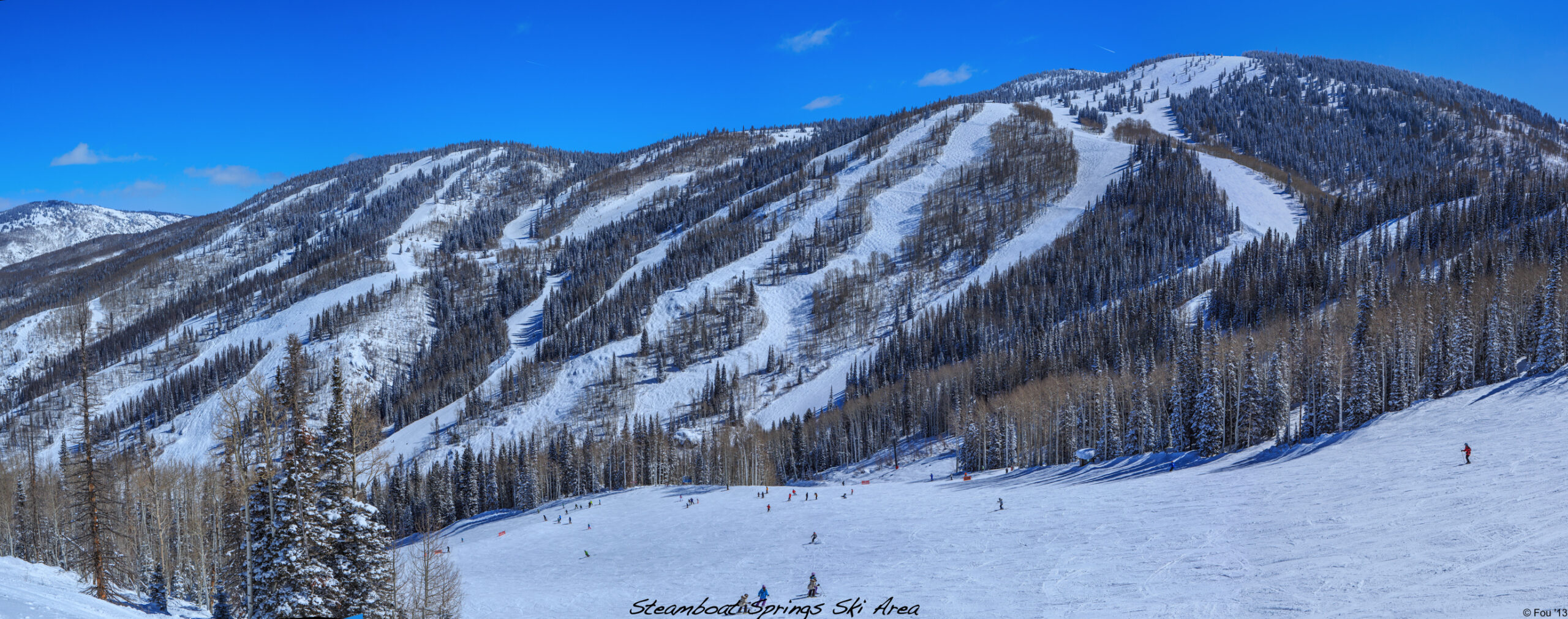 A panoramic view of a snowy mountain landscape at a ski resort. The image shows several ski slopes lined with snow-covered trees. Skiers and snowboarders are visible on the slopes, enjoying the clear, sunny day. The sky is bright blue with a few small clouds.