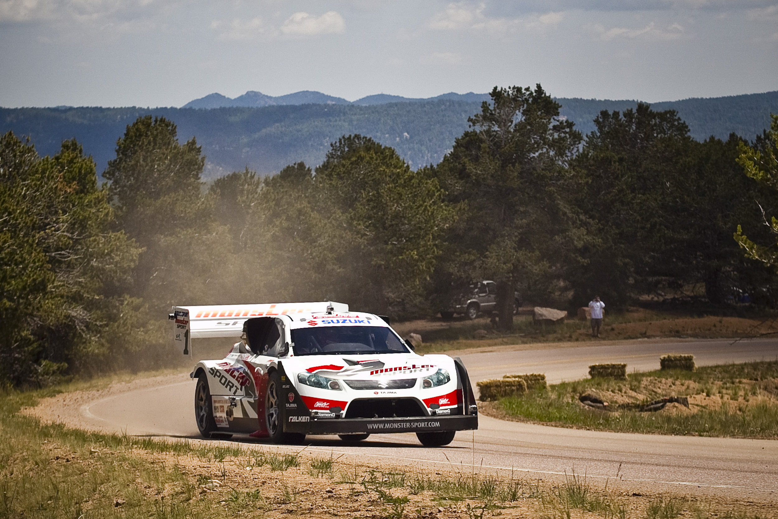 A race car is speeding around a curve on a mountain road, kicking up dust. The car is white with red and black accents and has various sponsor logos, including Suzuki and Monster. The background features trees and distant mountains under a partly cloudy sky. A person stands near the road, observing the scene.