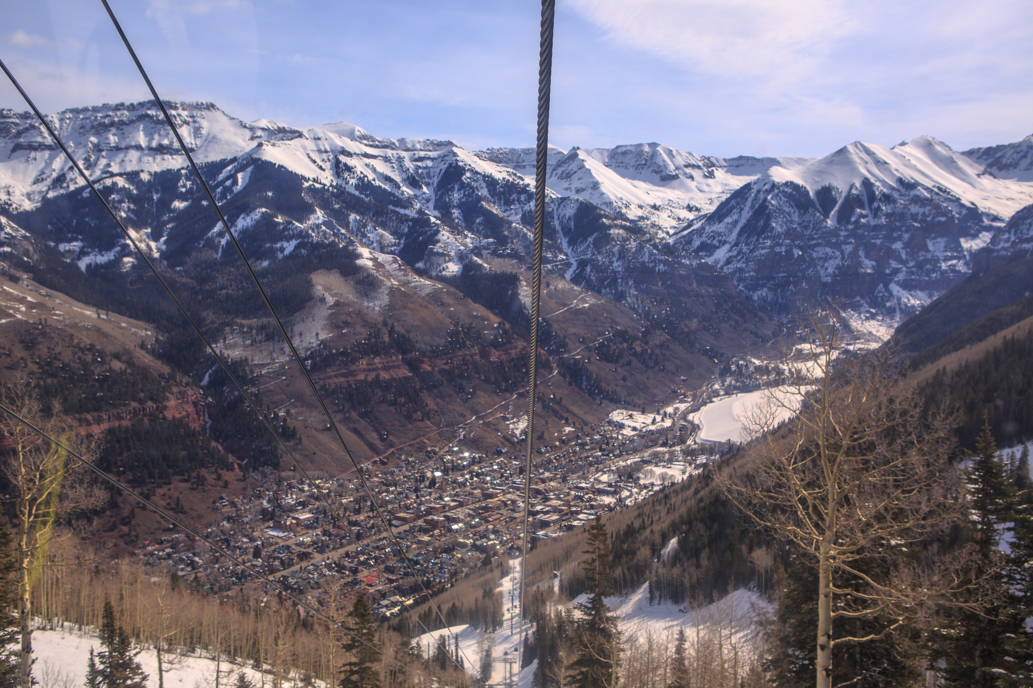 A scenic view of a mountainous landscape with snow-capped peaks in the background. In the foreground, there are ski lift cables and a few bare trees. Below, a small town is nestled in the valley, surrounded by forested hills and snow-covered areas. The sky is clear with a few clouds.