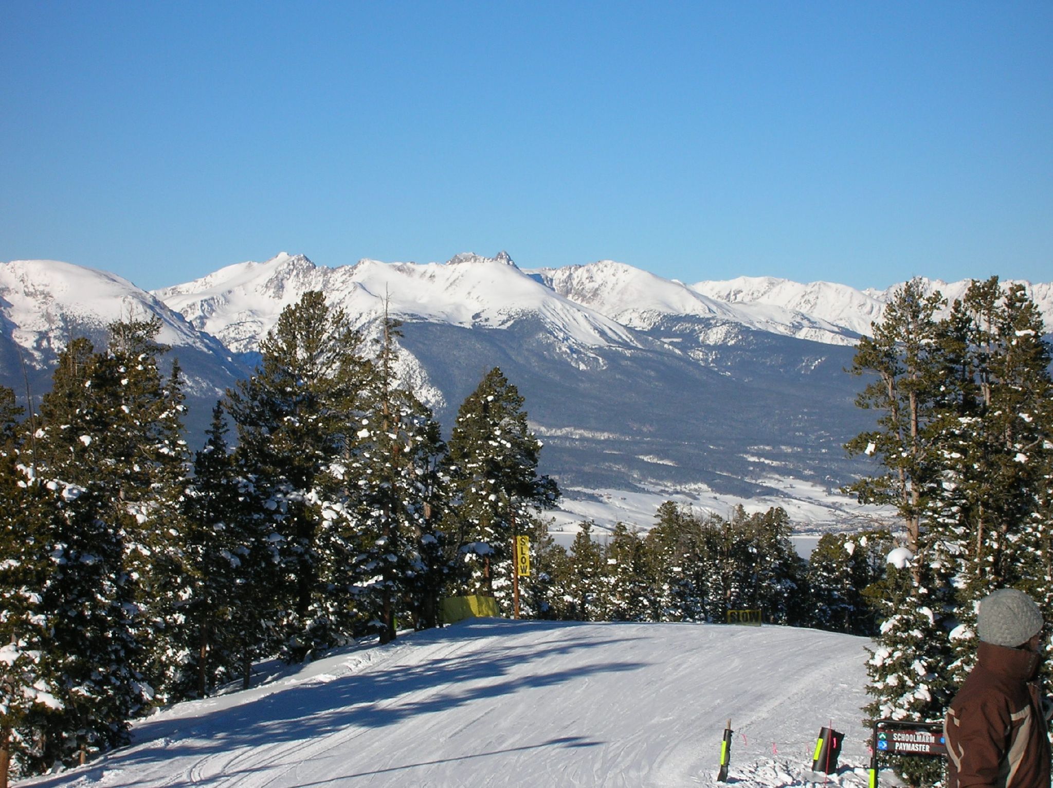 A snowy mountain landscape with a clear blue sky. In the foreground, there is a ski slope lined with snow-covered trees. Signs along the slope read "SLOW." In the background, a range of snow-capped mountains is visible. A person wearing a winter jacket and hat is standing on the right side of the image, looking towards the mountains.