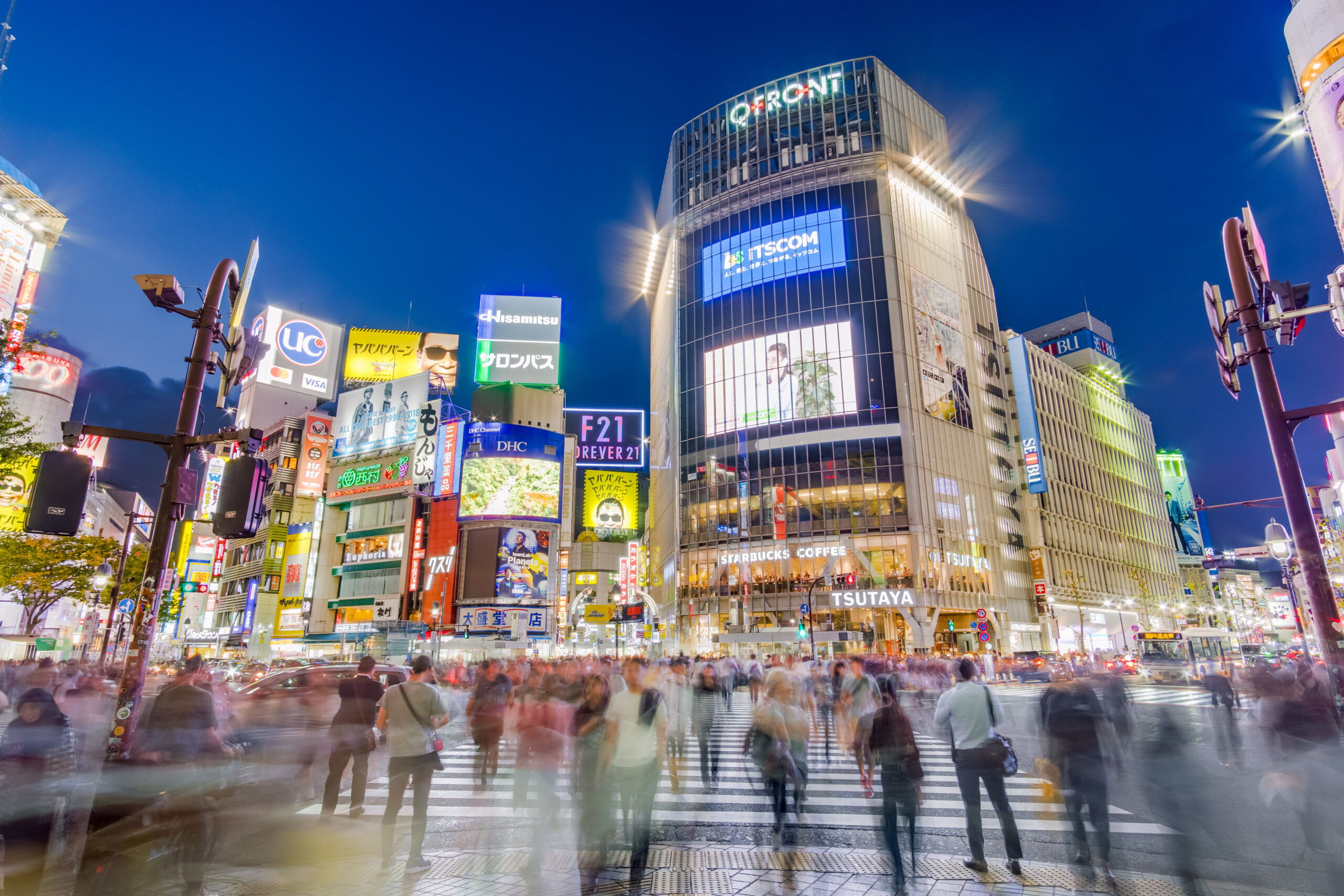 The image shows a bustling city scene at night, likely in Shibuya, Tokyo. Taken from Hachikō square. There are many people crossing the most famous scramble intersection in the world, with blurred motion indicating movement. Surrounding the intersection are tall buildings adorned with bright, colorful advertisements and billboards. The sky is clear and dark blue, suggesting evening time.