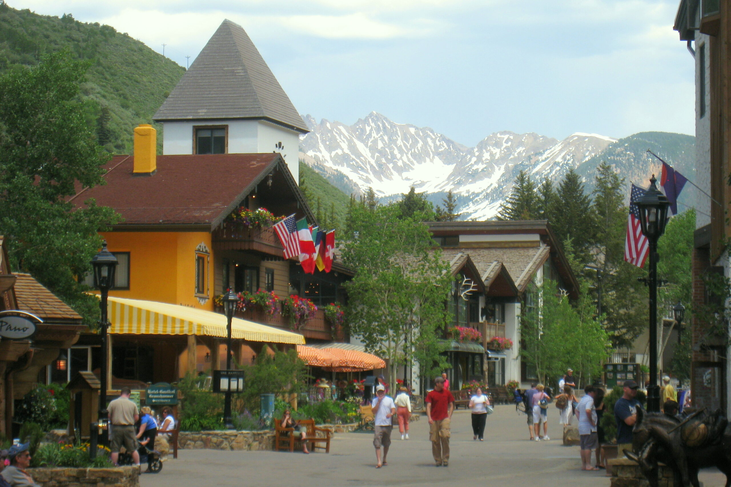 A picturesque village scene with a European-style architecture, featuring colorful buildings adorned with flowers and flags. People are walking along a pedestrian street, and there are outdoor seating areas with umbrellas. In the background, snow-capped mountains rise against a partly cloudy sky, surrounded by lush green trees.