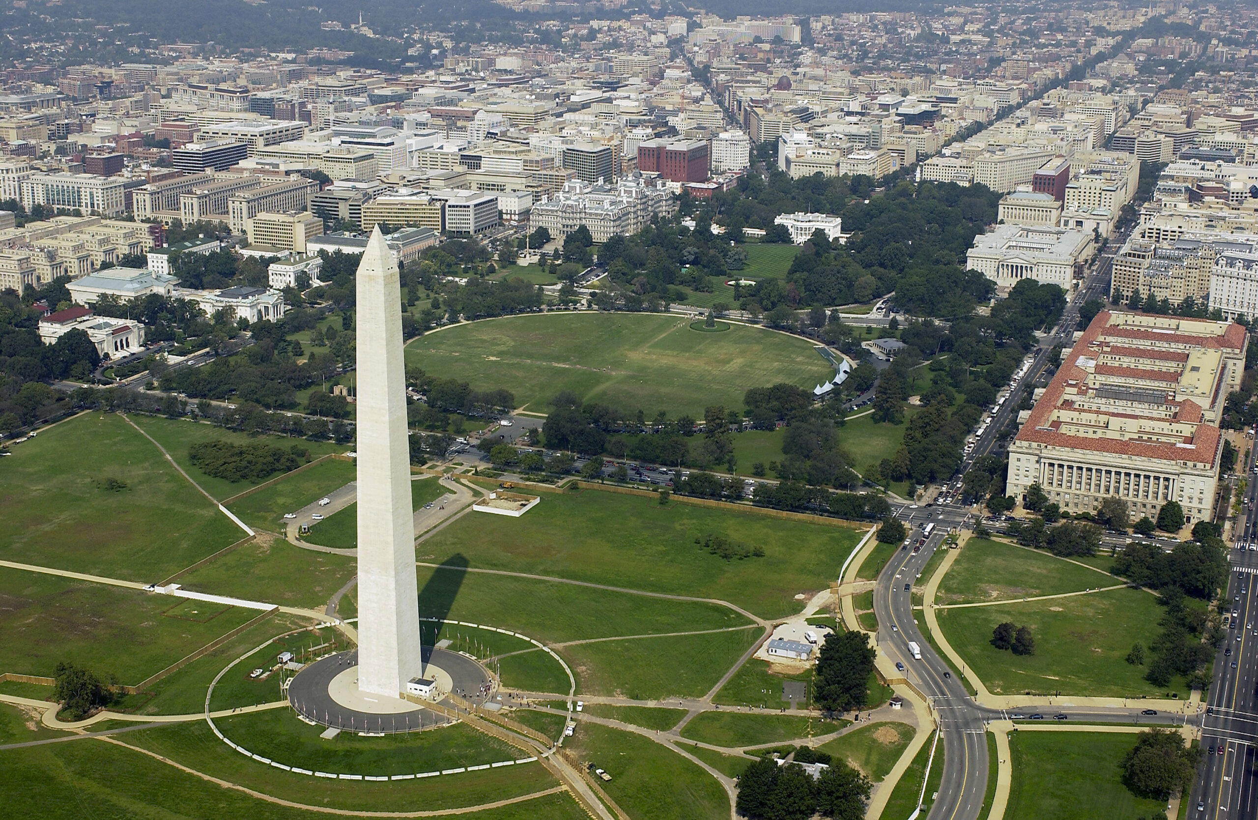 Aerial view of the Washington Monument, a tall, white obelisk surrounded by green lawns and pathways. The surrounding area includes numerous buildings and tree-lined streets, with a cityscape in the background.