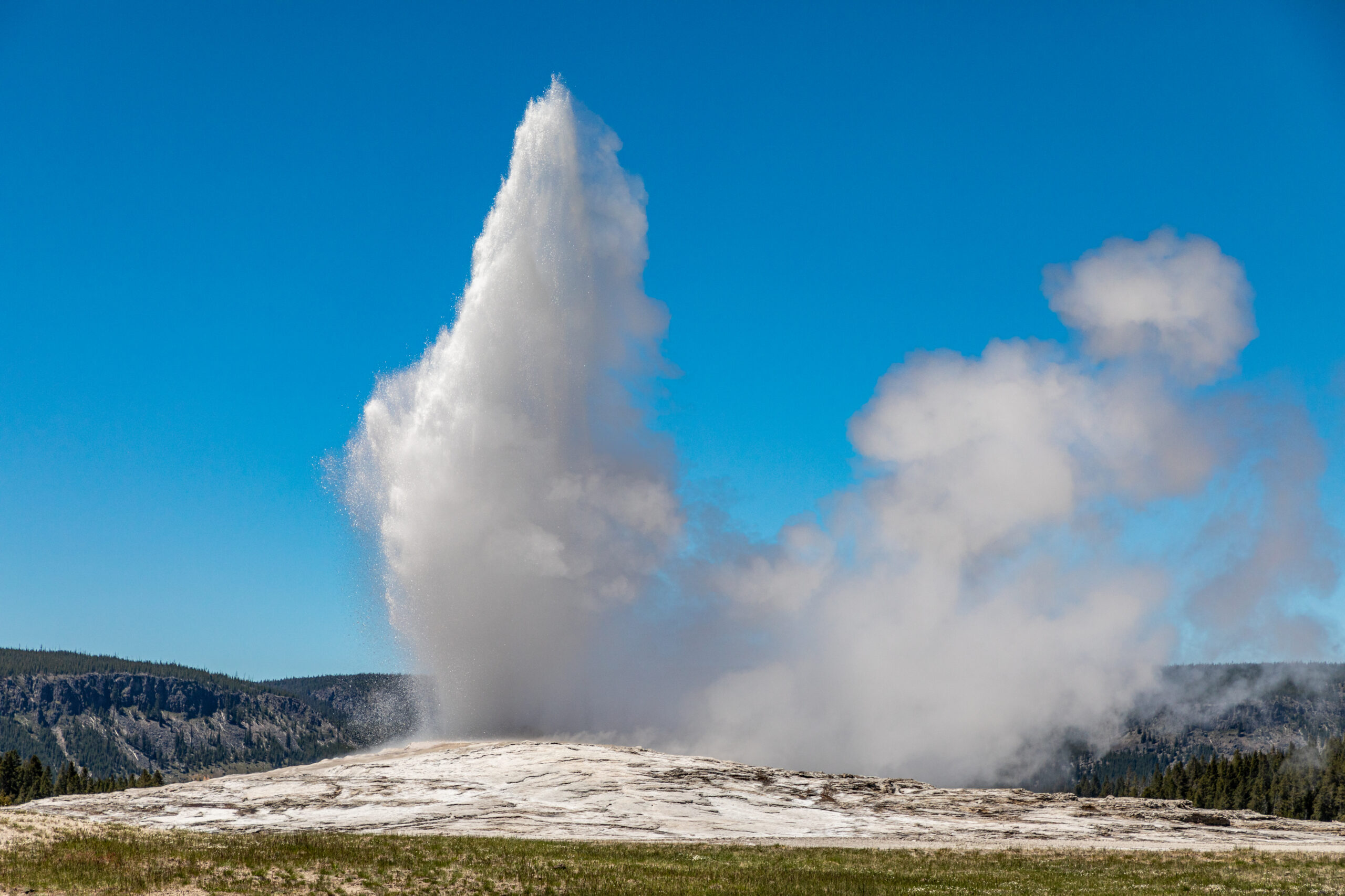 The image shows the famous Old Faithful Geyser at Yellowstone National Park. A large geyser is seen erupting under a clear blue sky. The geyser, surrounded by mist and water vapor, shoots a powerful stream of water high into the air. The landscape around the geyser includes a rocky surface, with distant forests and mountainous terrain visible in the background. 