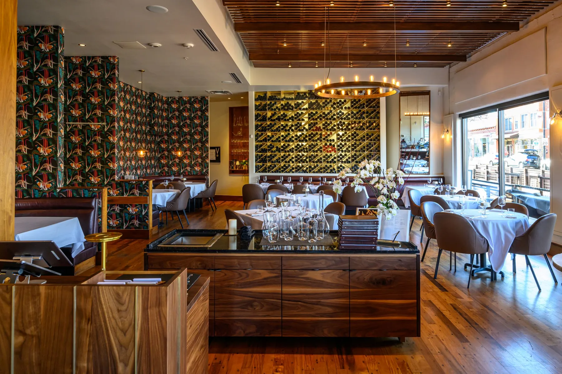 The image shows the interior of a stylish restaurant. The space features wooden floors and a ceiling with wooden slats. There are tables set with white tablecloths and brown chairs. A large wine rack filled with bottles is visible on one wall. The walls have a decorative floral pattern, and there are large windows allowing natural light to enter. A chandelier with circular lights hangs from the ceiling, and a wooden reception desk is in the foreground.