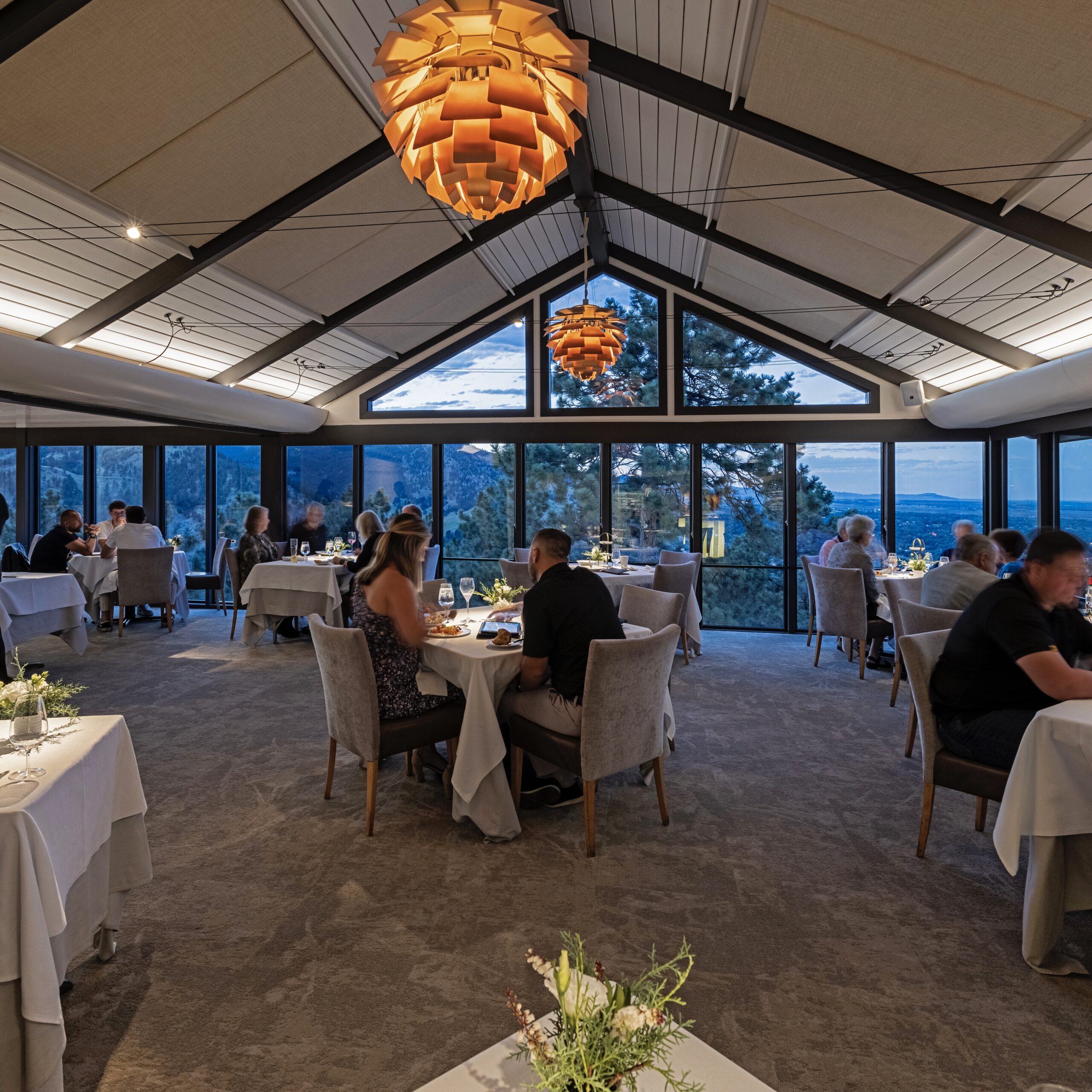 The image shows a modern restaurant interior with large windows offering a scenic view of a mountainous landscape. Several tables are set with white tablecloths, and people are dining in a relaxed atmosphere. The ceiling features unique, warm-toned pendant lights, and the room has a high, sloped ceiling with exposed beams. The setting appears to be during the evening, with a soft, ambient light.